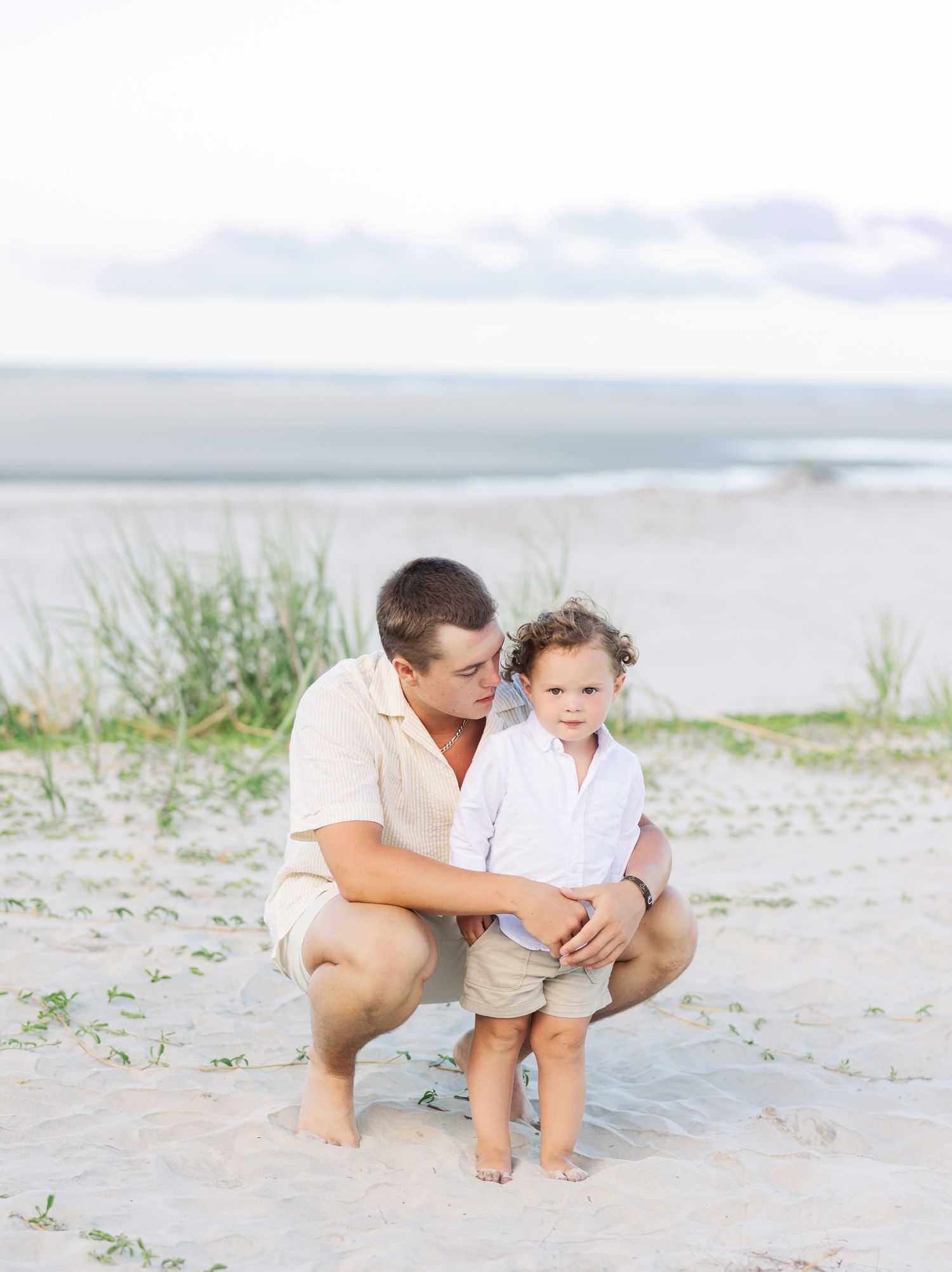 A tender beach scene with an adult and small child sharing a quiet moment in the sand near the ocean.
