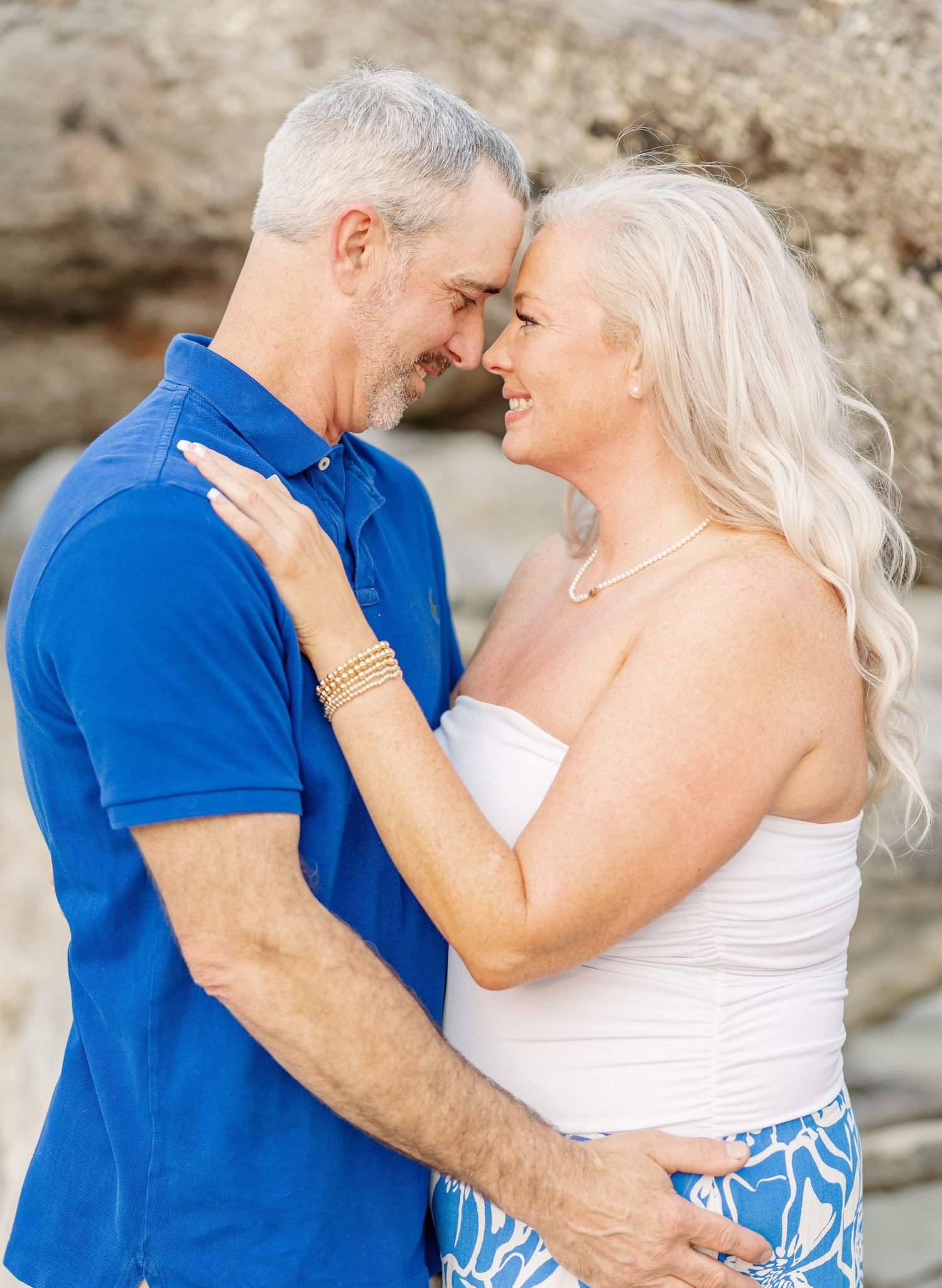 Romantic couple in blue and white embracing and sharing intimate moments on a beach with rocky backdrop.