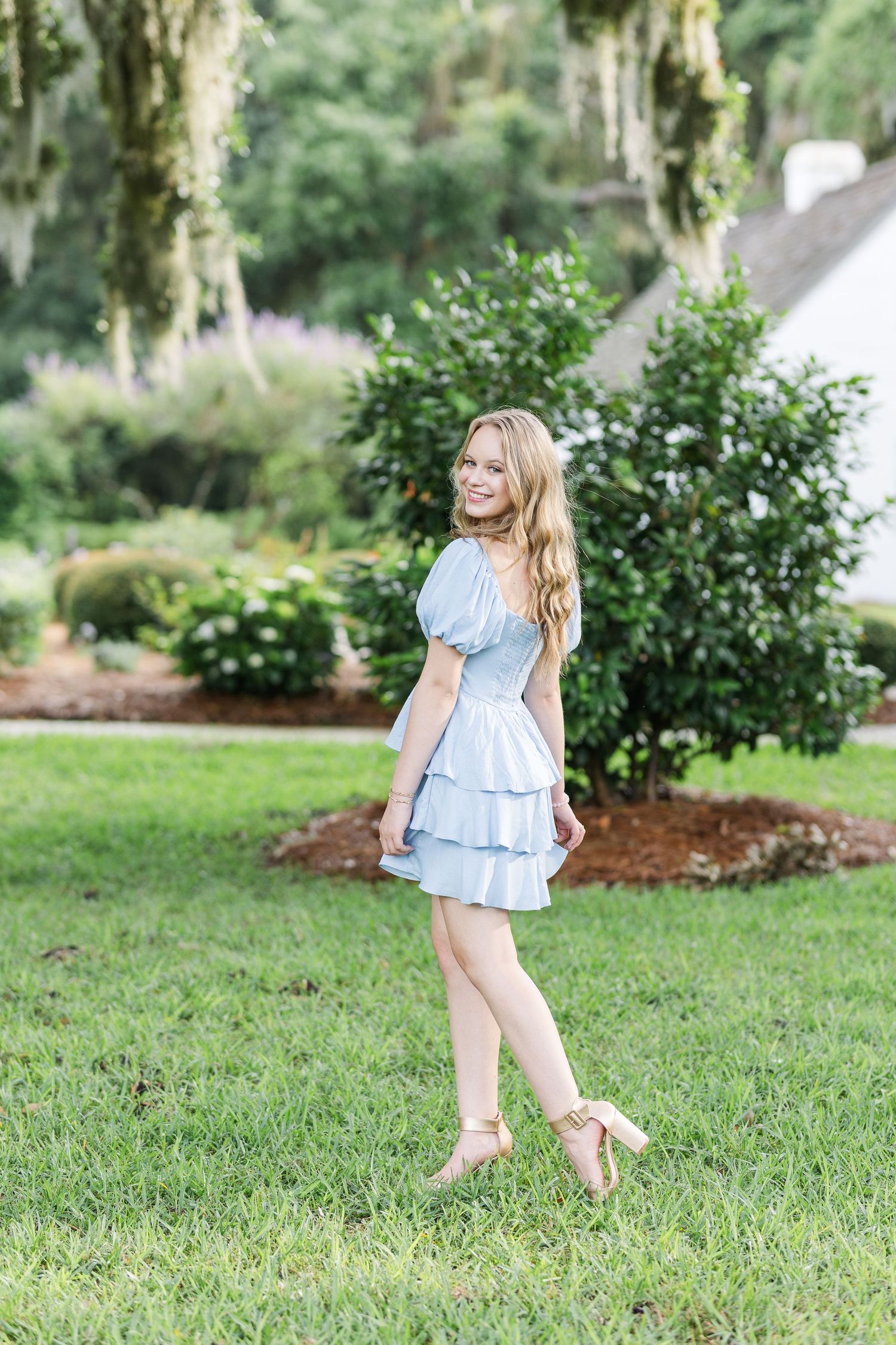 A woman in a blue tiered dress and heels stands in a lush garden setting.