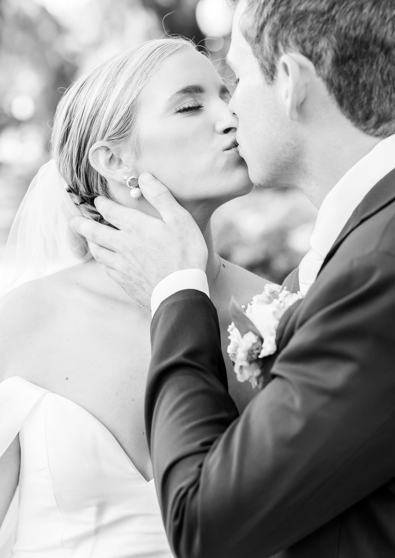 A romantic black and white wedding photograph capturing a tender kiss moment between the bride and groom outdoors.