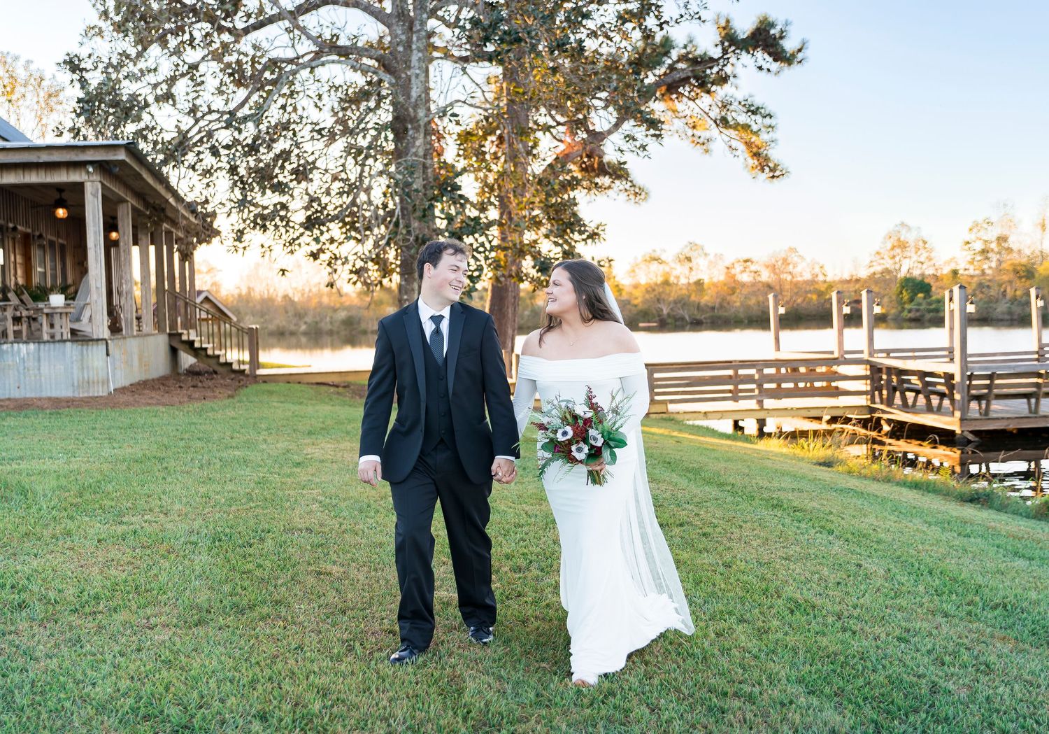 Couple poses together on manicured lawn with lake and dock in background during sunset.