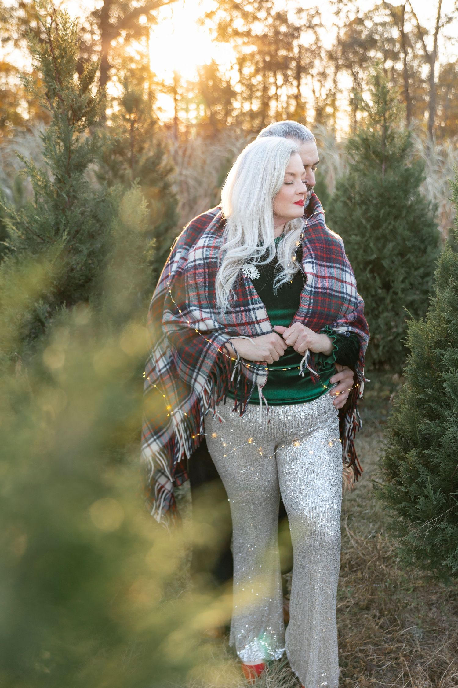 Person in sparkly pants and plaid wrap poses thoughtfully among Christmas trees at sunset.