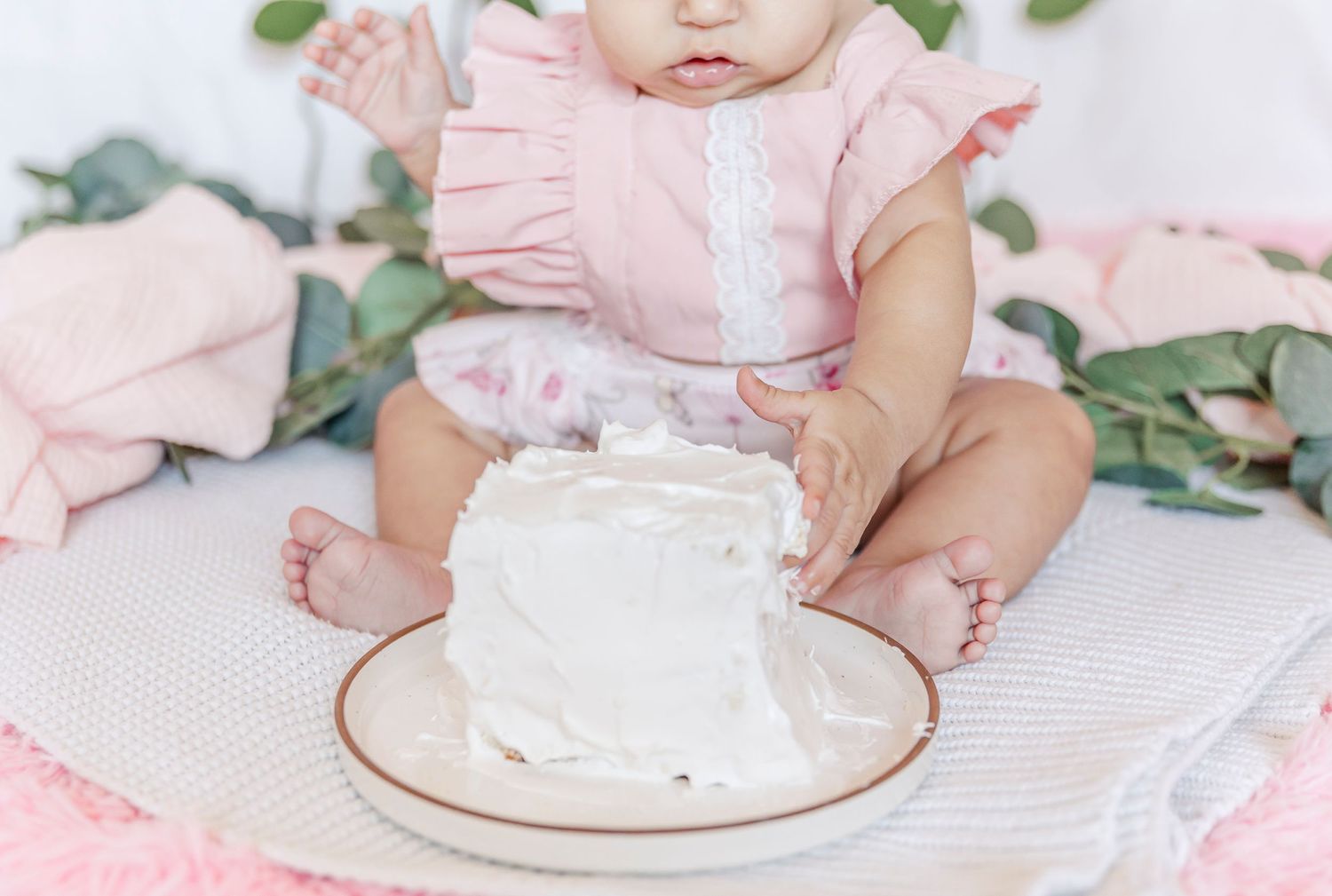 Baby in pink dress looking at small white cake on plate with floral background.