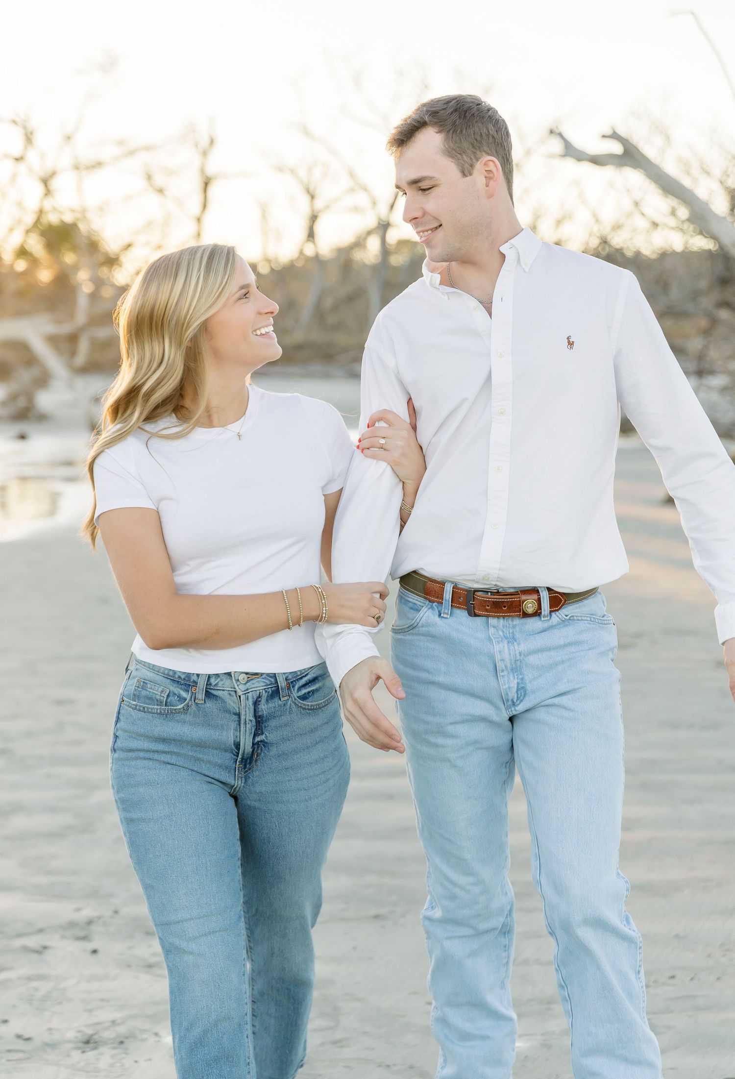 A couple in coordinated white and denim outfits walks together on beach.