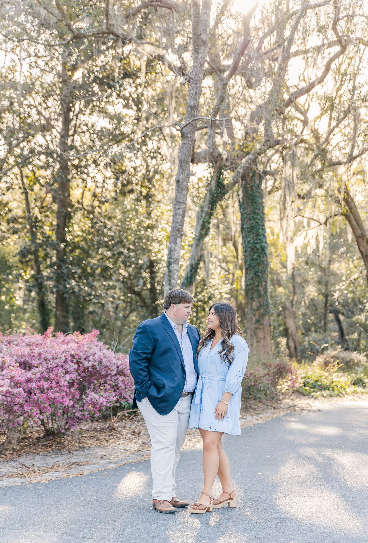 A romantic couple stands together on a tree-lined path with pink flowering bushes in soft natural lighting.