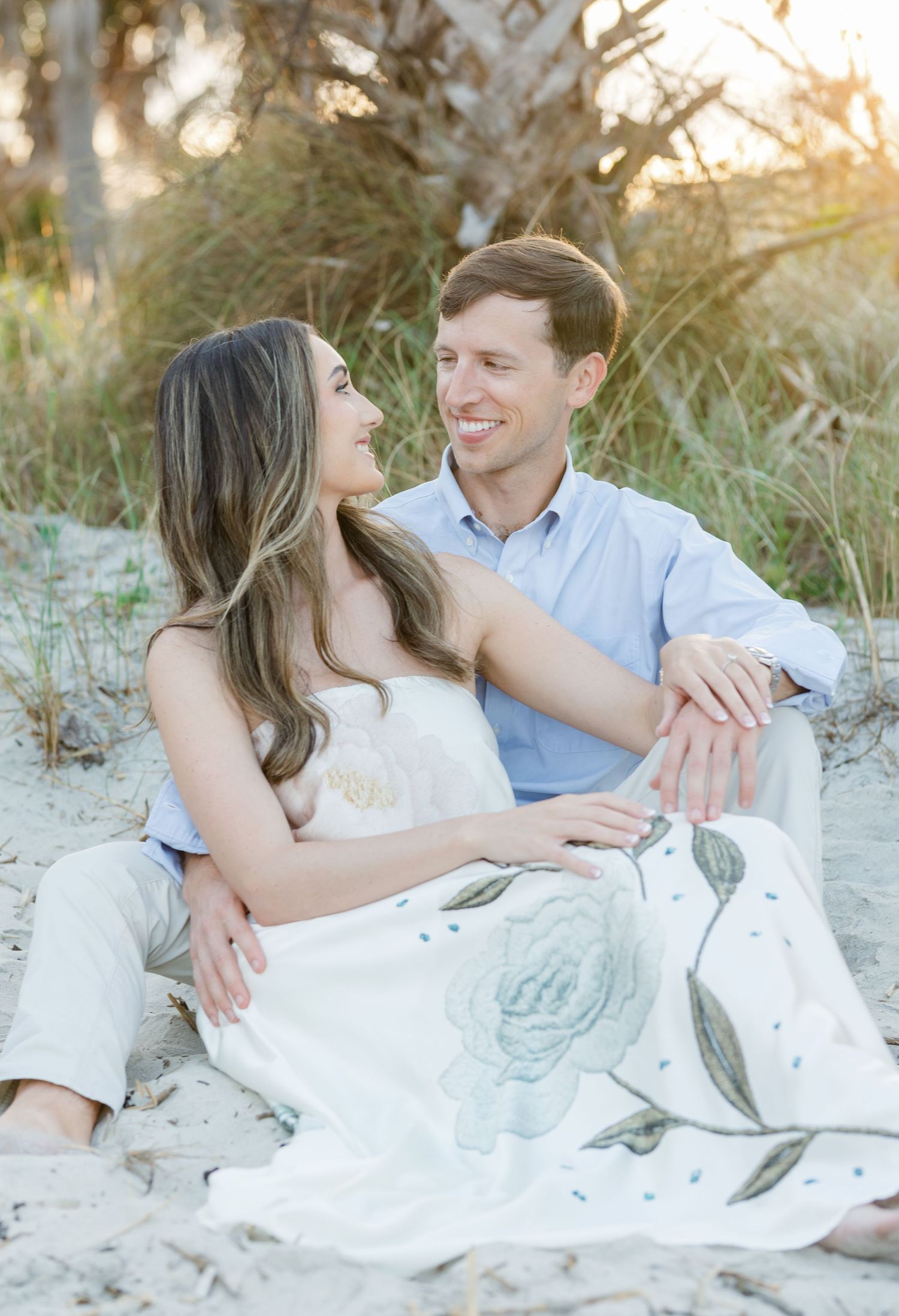 Couple sitting together on sandy beach dunes, smiling at each other, woman in floral skirt and man in blue shirt.