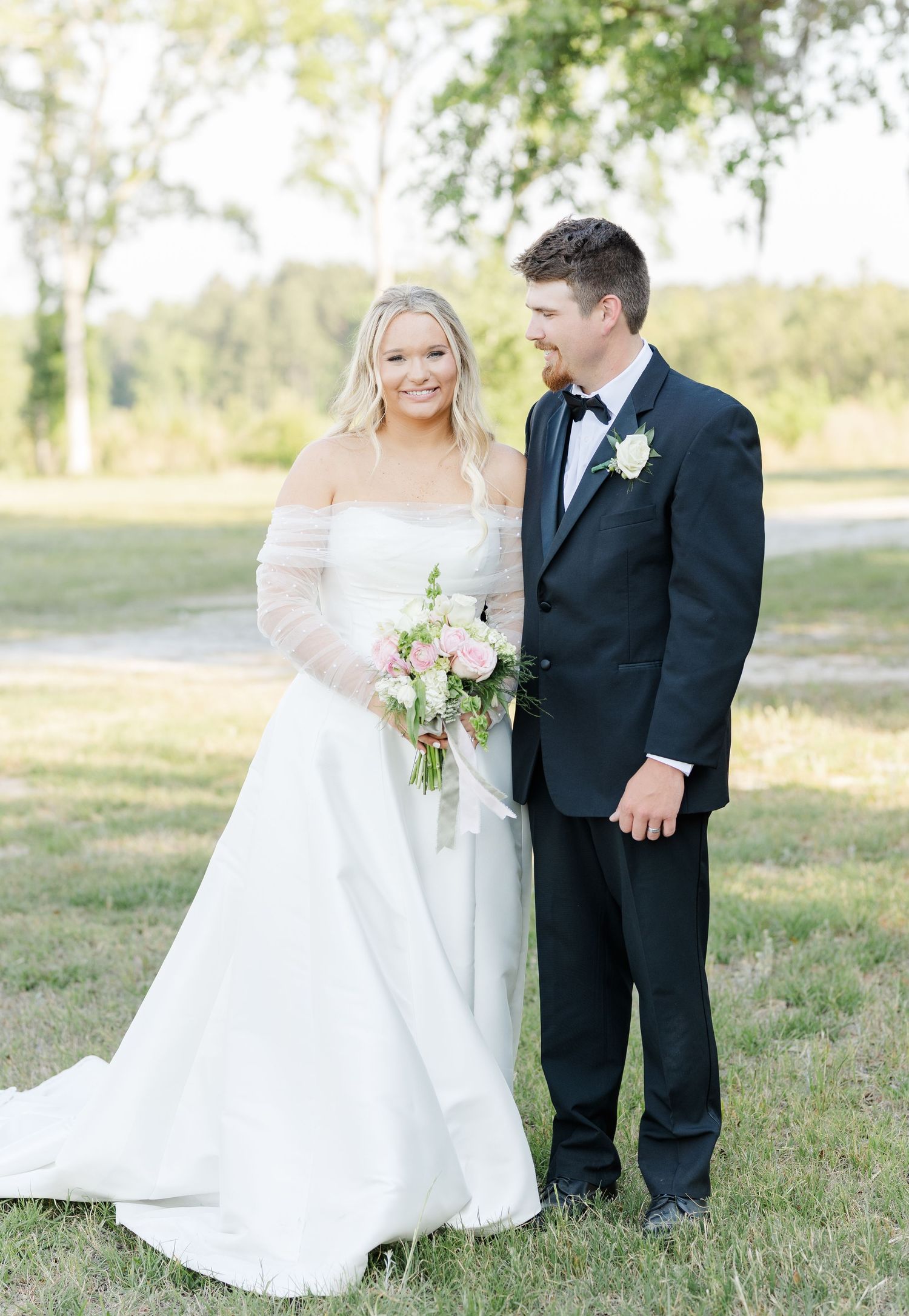 Bride in off-shoulder gown with sheer sleeves holding pink bouquet beside groom in black tux.