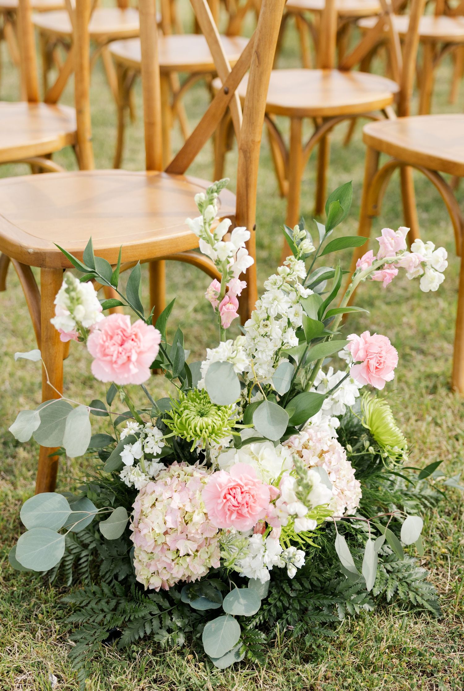 Pink carnations, hydrangeas, and white flowers ceremony aisle marker beside wooden cross-back chairs.