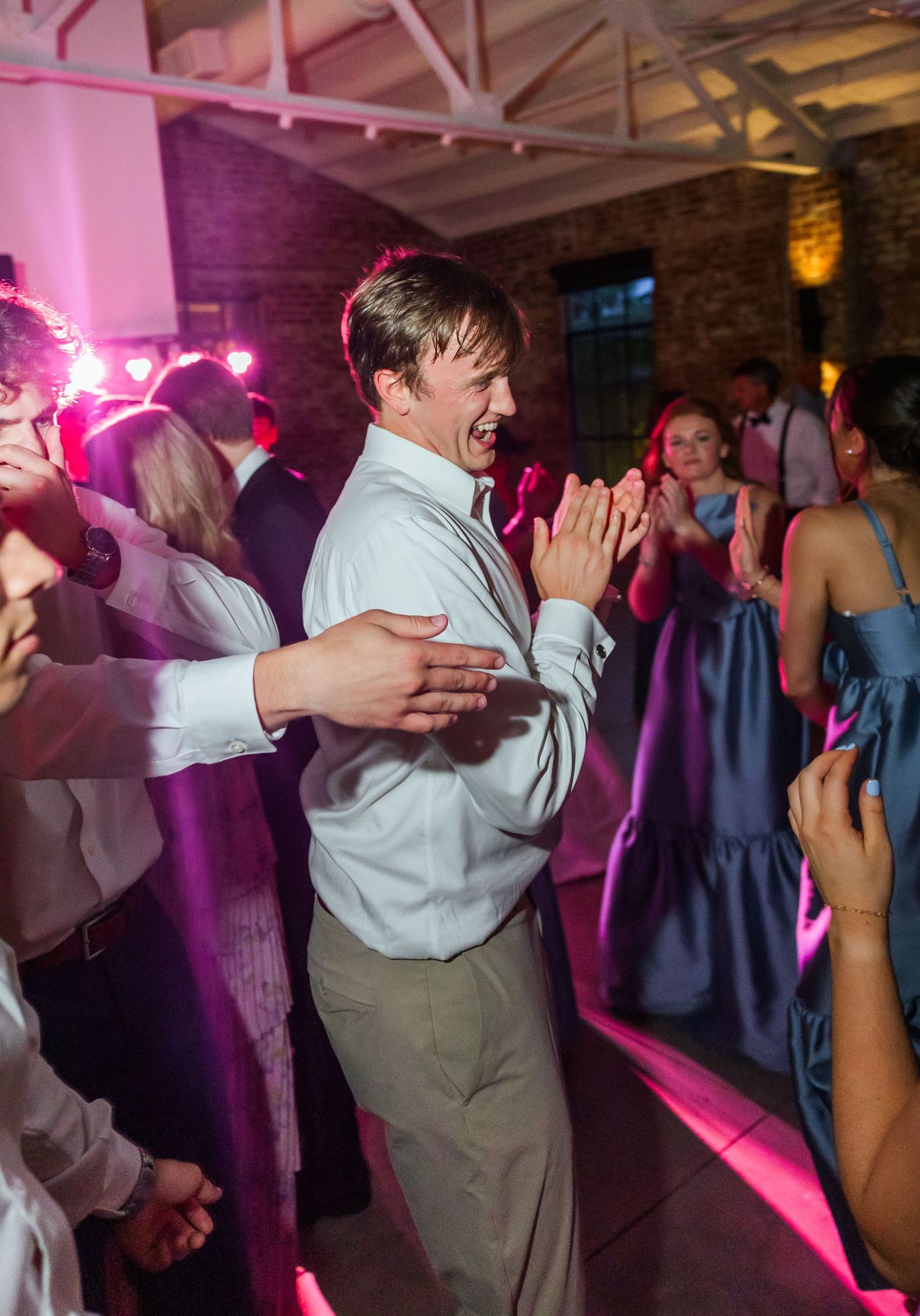 A young man in a white shirt dances energetically at a lively wedding reception with pink lighting.