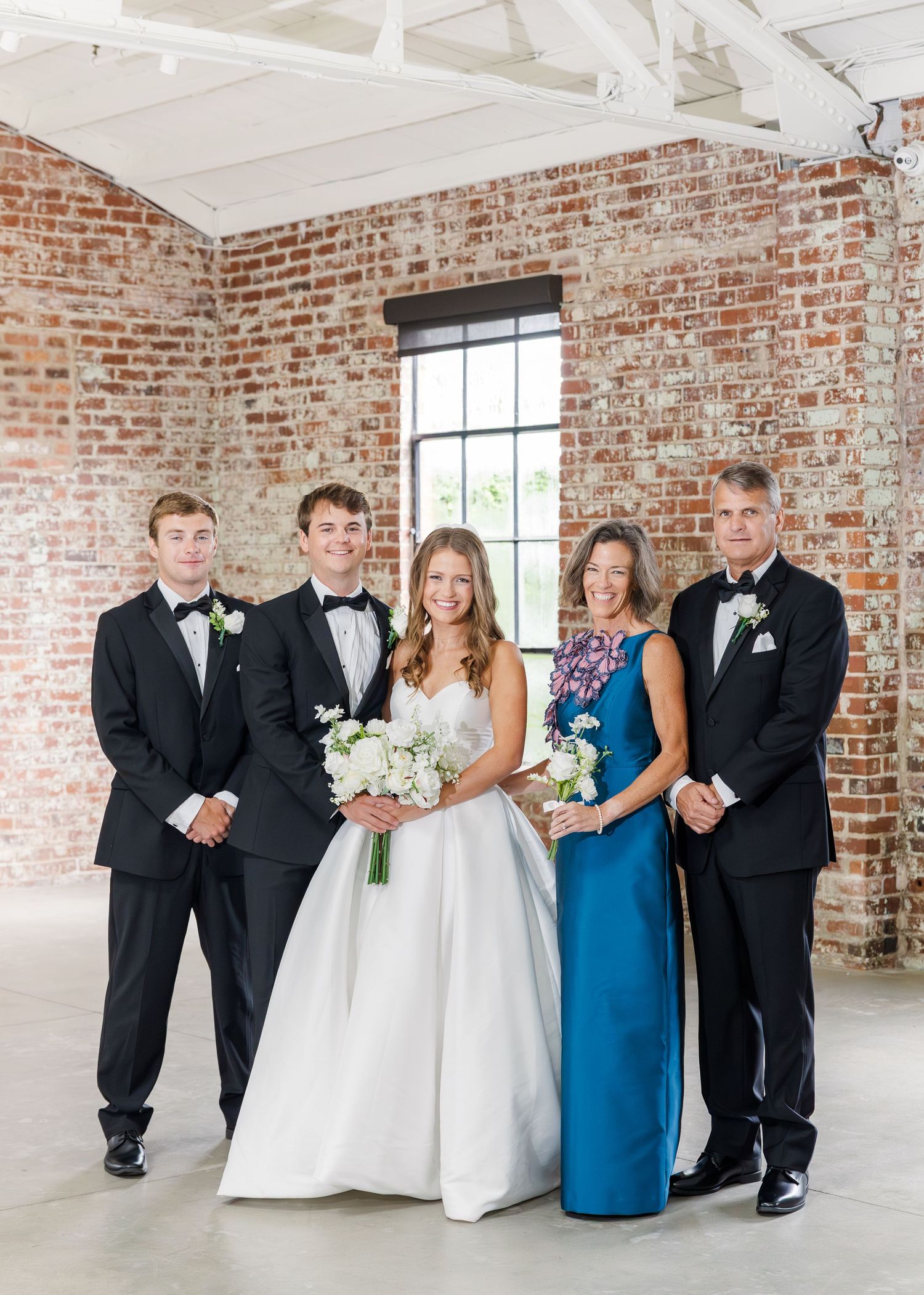 Family portrait with bride in white gown and groom in tuxedo flanked by relatives in elegant attire at brick wall venue.
