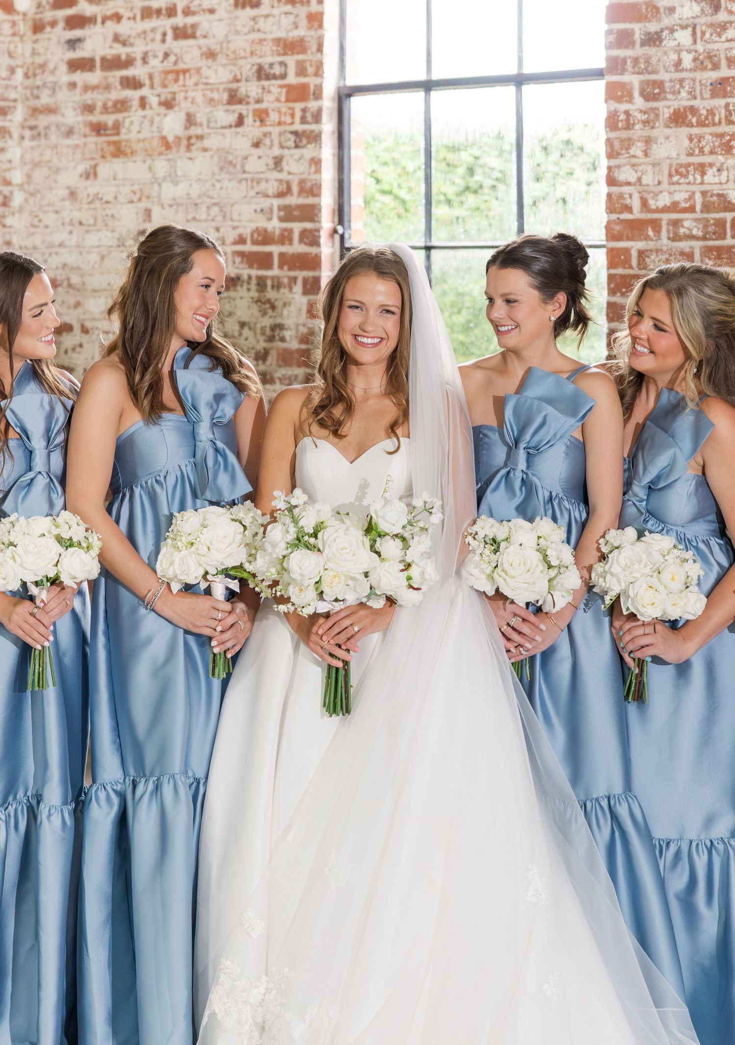 Smiling bride in white gown with veil surrounded by bridesmaids in dusty blue dresses holding white bouquets.