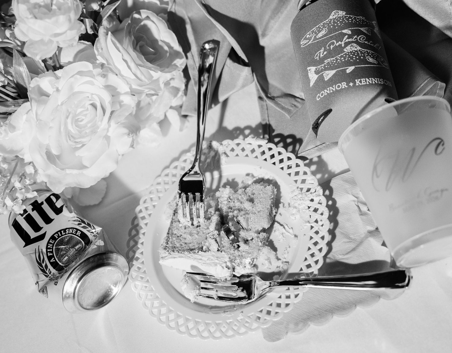 Black and white photo of wedding reception table with cake slice, Miller Lite can, wine bottle, and floral arrangement.