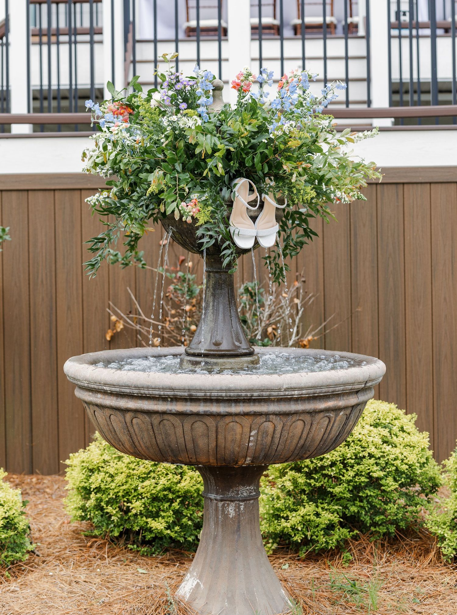 White bridal heels displayed on a stone garden fountain surrounded by lush floral arrangements with blue and coral blooms.