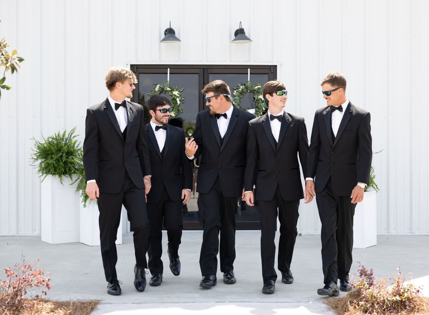 Five groomsmen in black tuxedos and bow ties walking together outside a modern white wedding venue.