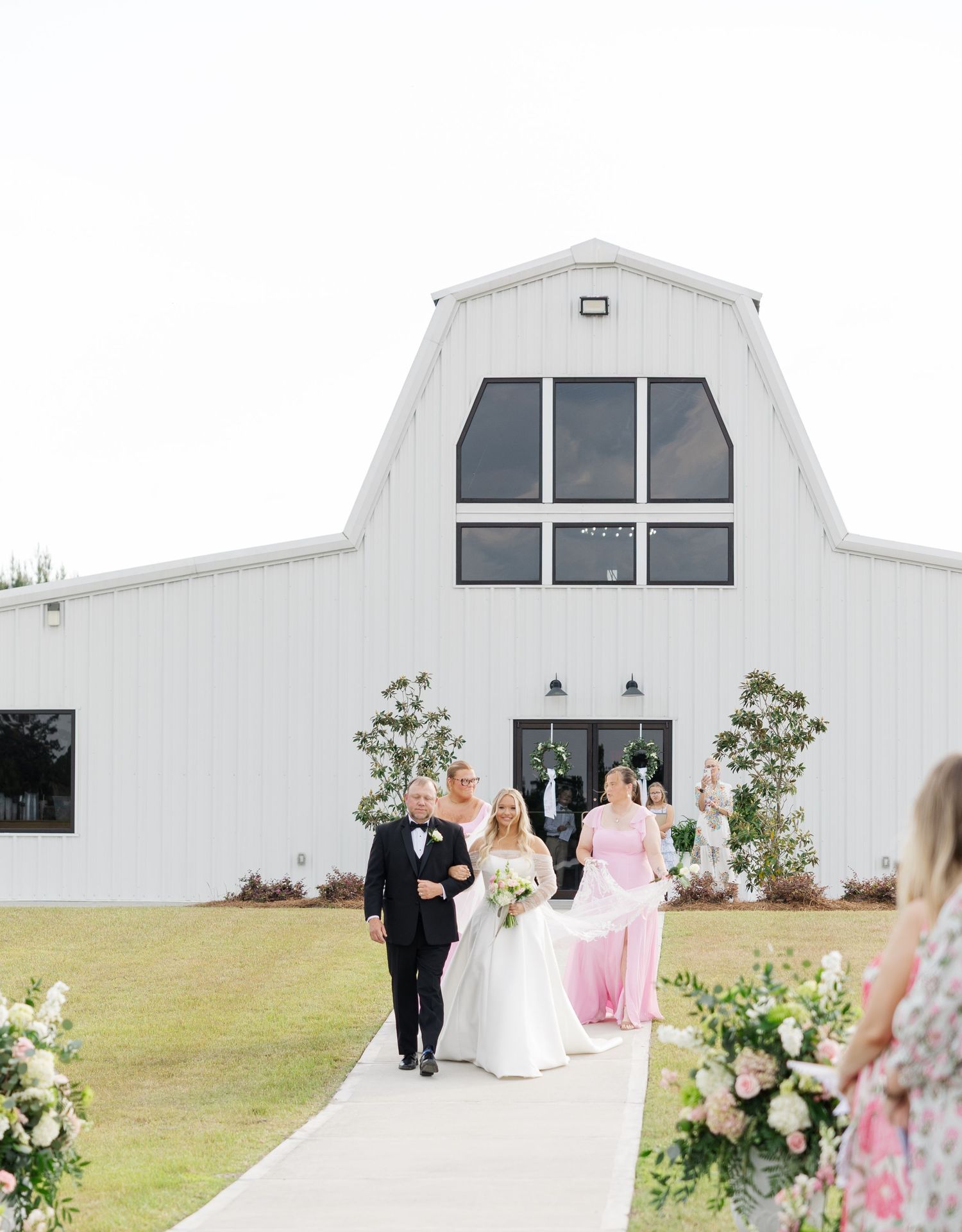 Bride in white gown escorted down aisle by father at white barn wedding venue.