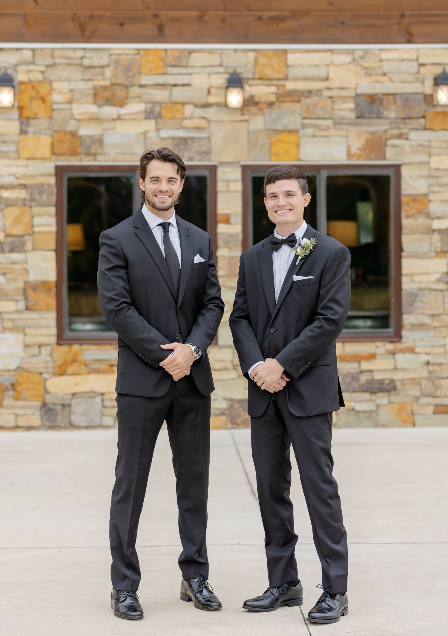 Two groomsmen in dark suits pose against stone wall with lanterns.