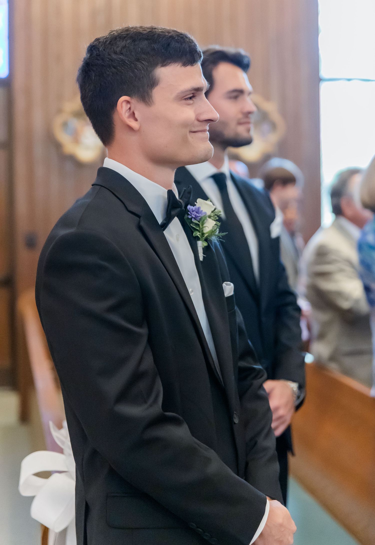 Wedding ceremony attendants in formal attire sit in wooden church pews.