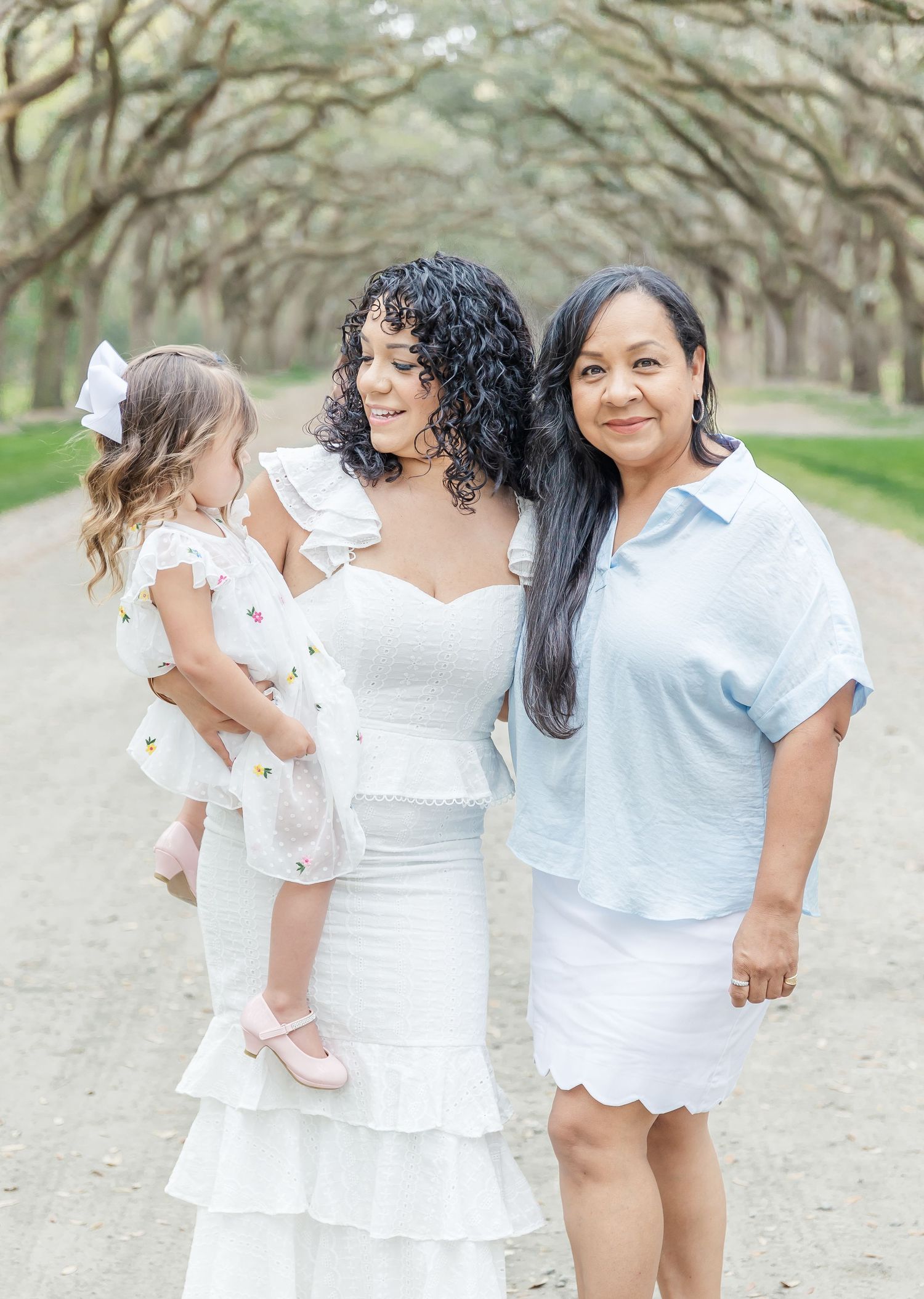 Three generations pose together on a tree-lined dirt path in coordinating white and light blue outfits.