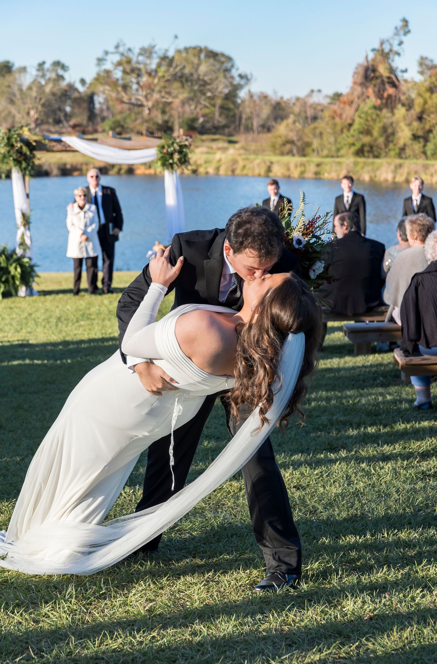 Romantic dip kiss between newlyweds during lakeside wedding ceremony.
