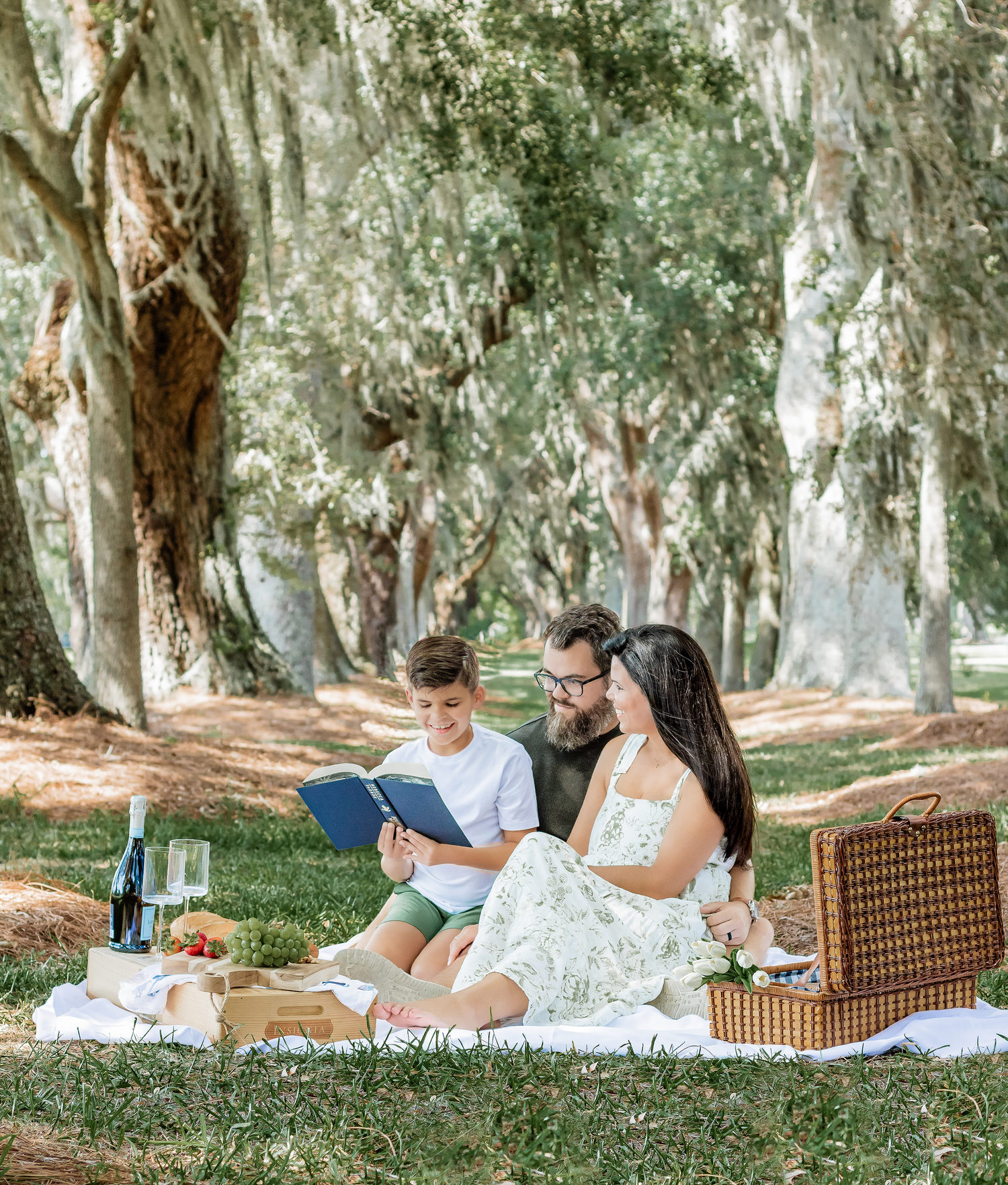 A couple enjoys a romantic picnic under moss-draped trees with a wicker basket and blanket.