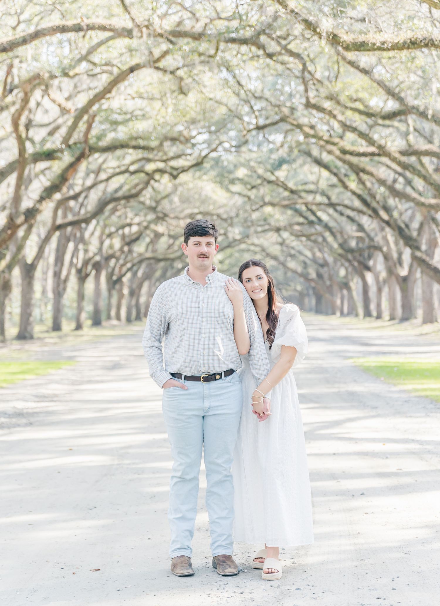 A romantic couple poses under a canopy of oak trees on a dreamy pathway wearing coordinated light-colored outfits.