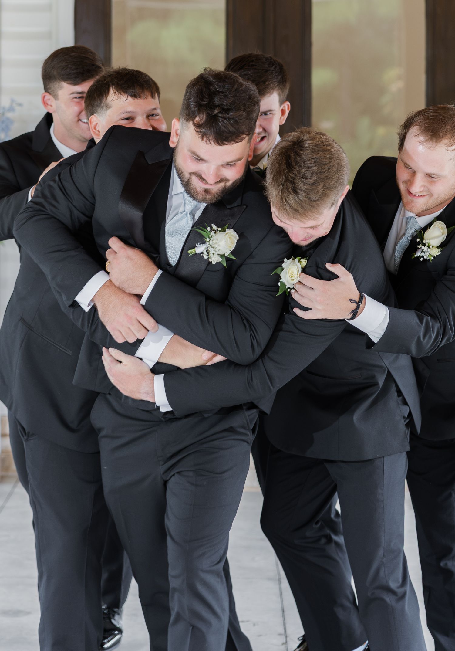 Groom laughing as groomsmen in dark suits give him a group hug before wedding ceremony.