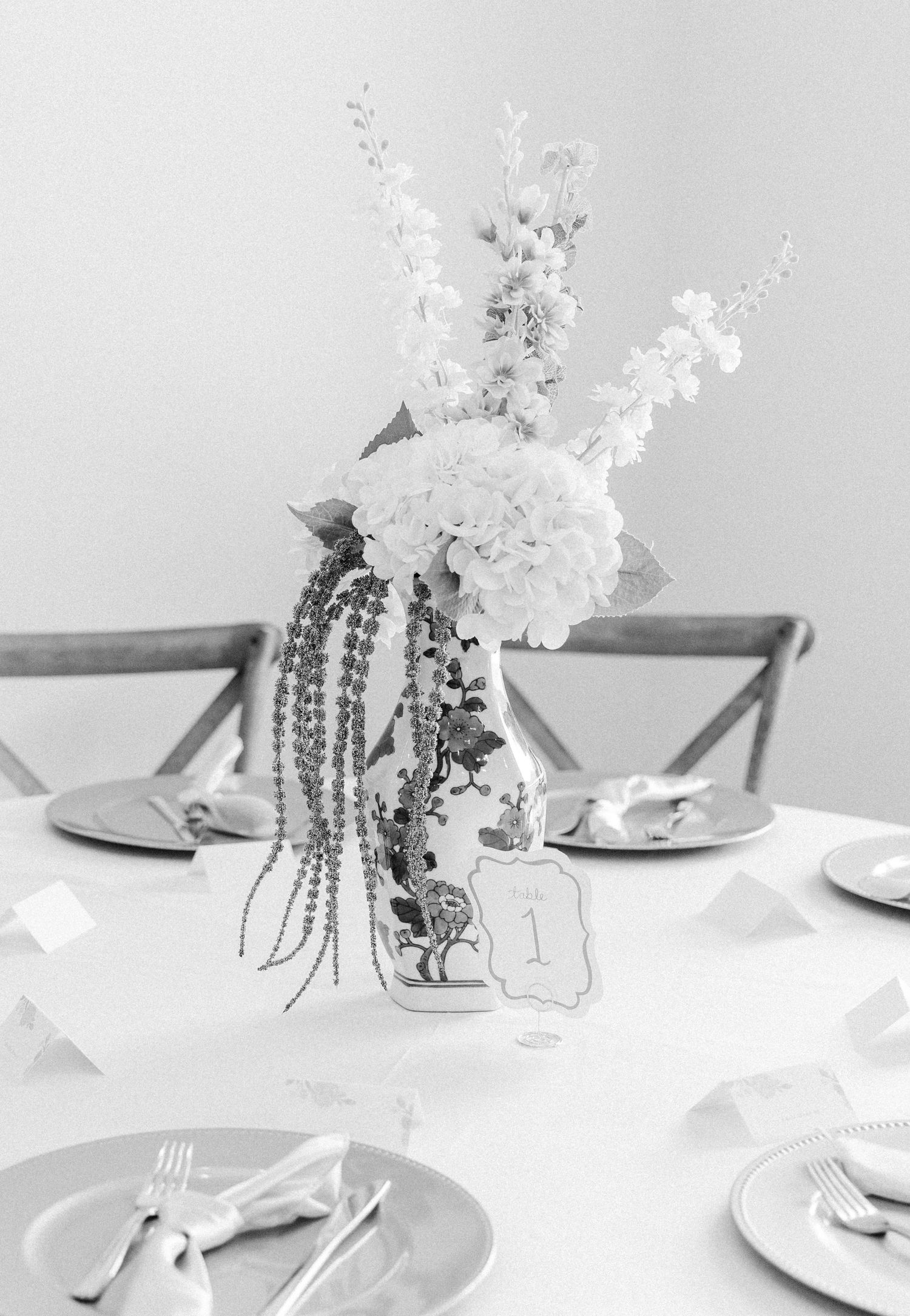 Black and white photo of elegant wedding table centerpiece with white flowers in decorative vase, table number one visible.