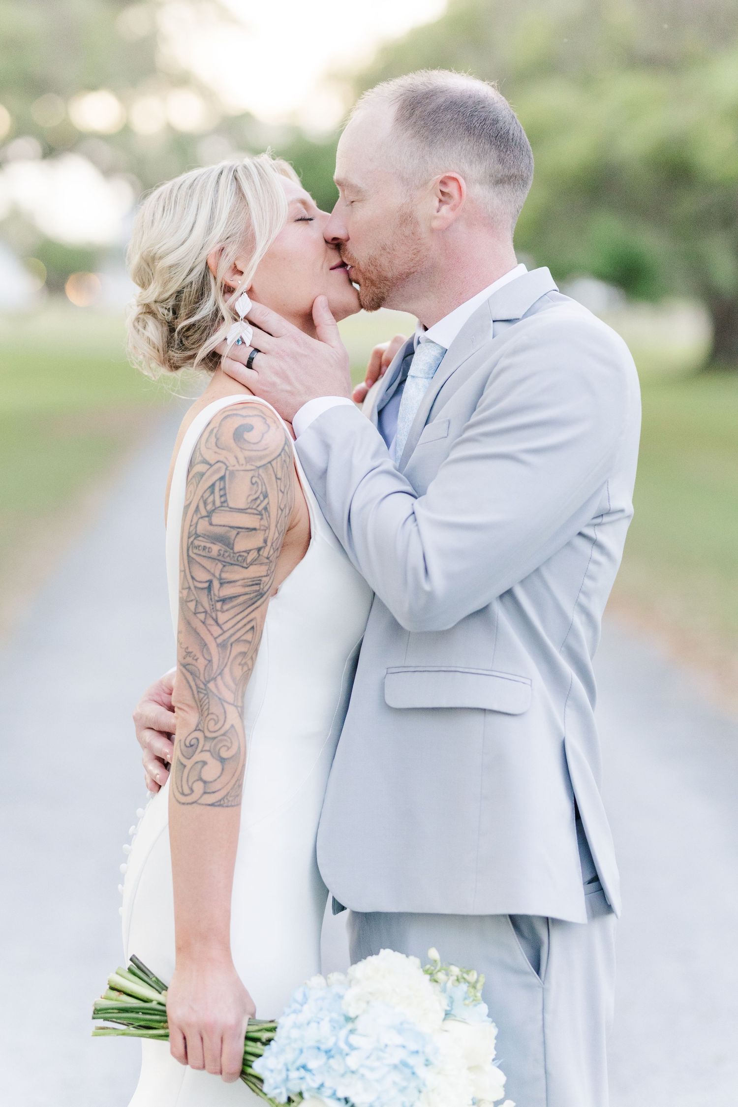 Bride with sleeve tattoo and groom in light blue suit sharing first kiss outdoors.