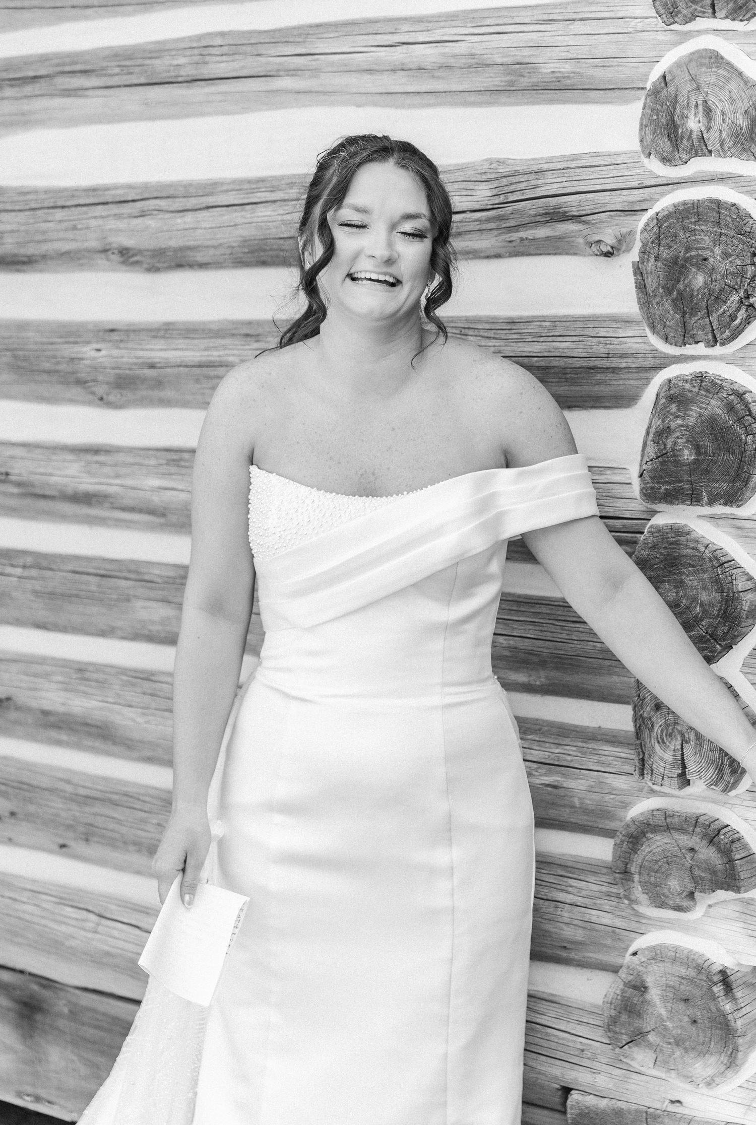 Bride in off-shoulder white gown laughing joyfully against rustic log wall in black and white wedding photo.