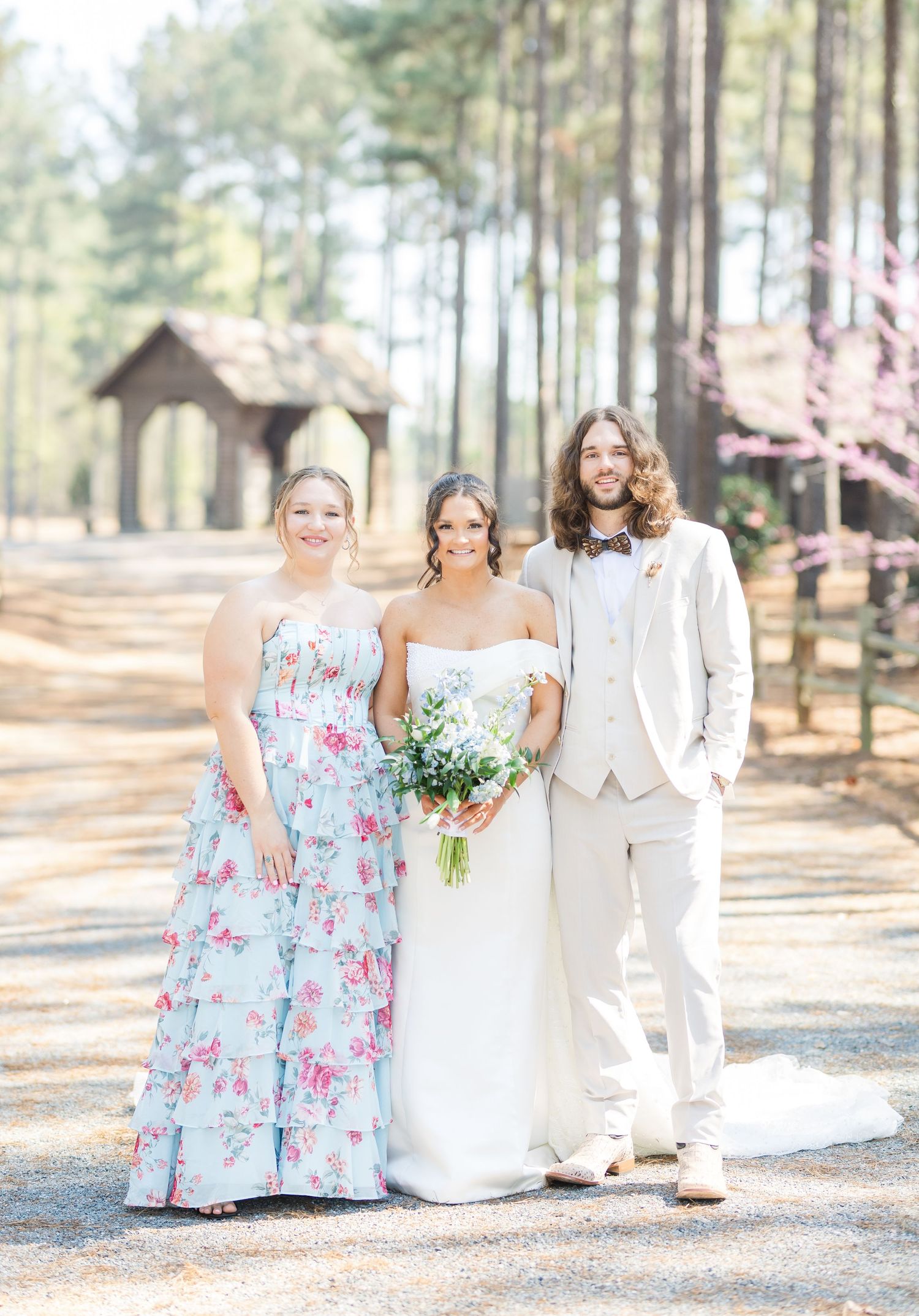 Bride and groom pose with a bridesmaid in a floral dress in a pine forest wedding setting.