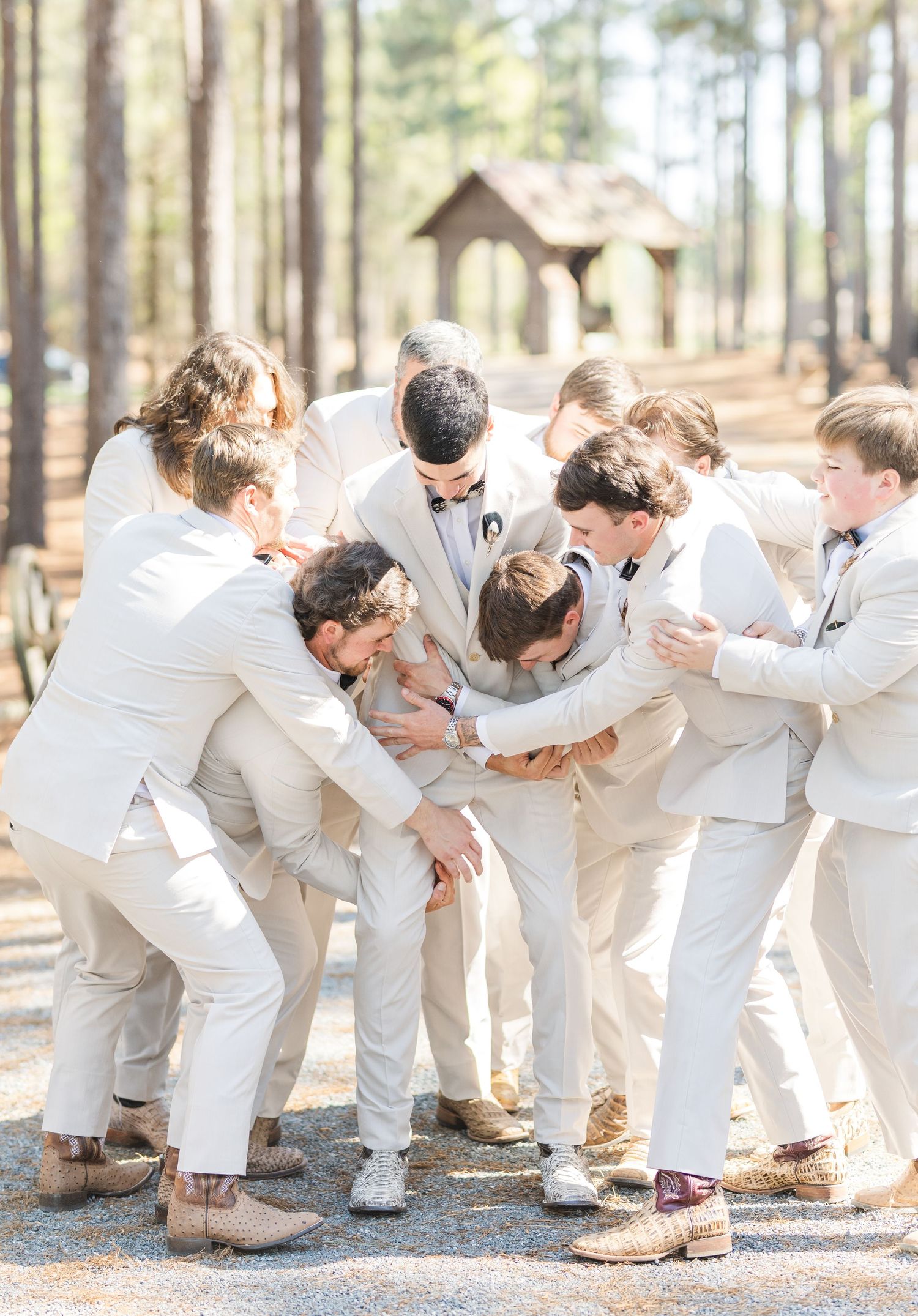 Groomsmen in tan suits playfully huddle around the groom on a gravel path near a rustic outdoor wedding chapel.