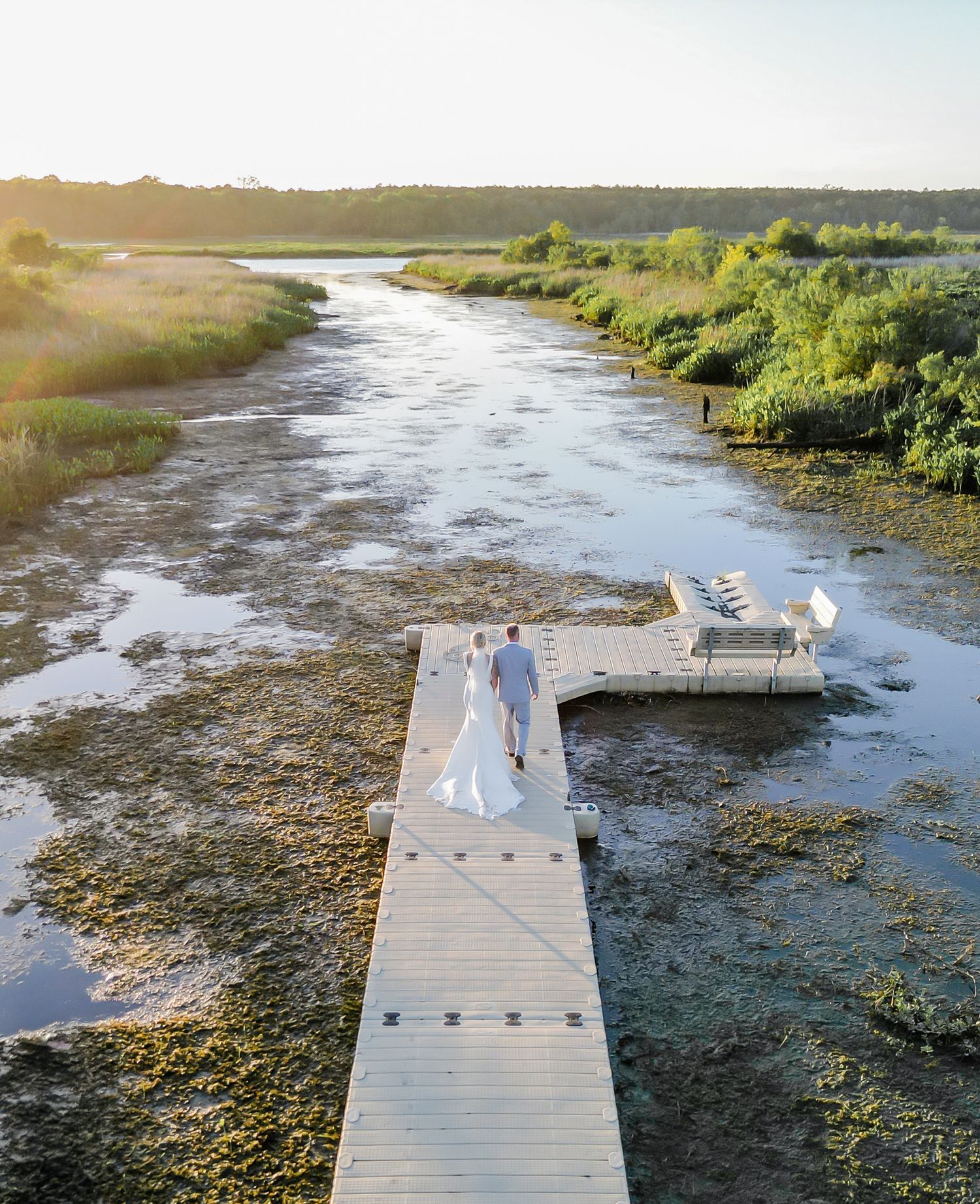 Aerial drone view of bride and groom walking on wooden dock over scenic coastal marshland at sunset.