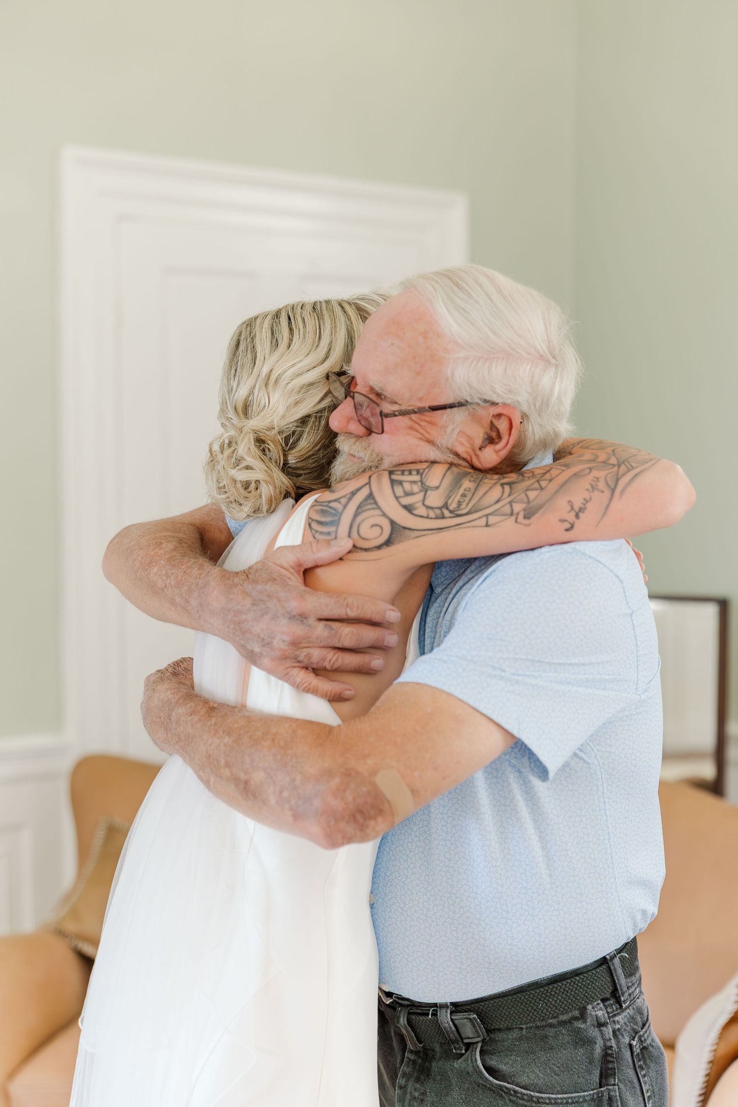 Emotional bride with sleeve tattoo shares tender hug with elderly father before wedding.