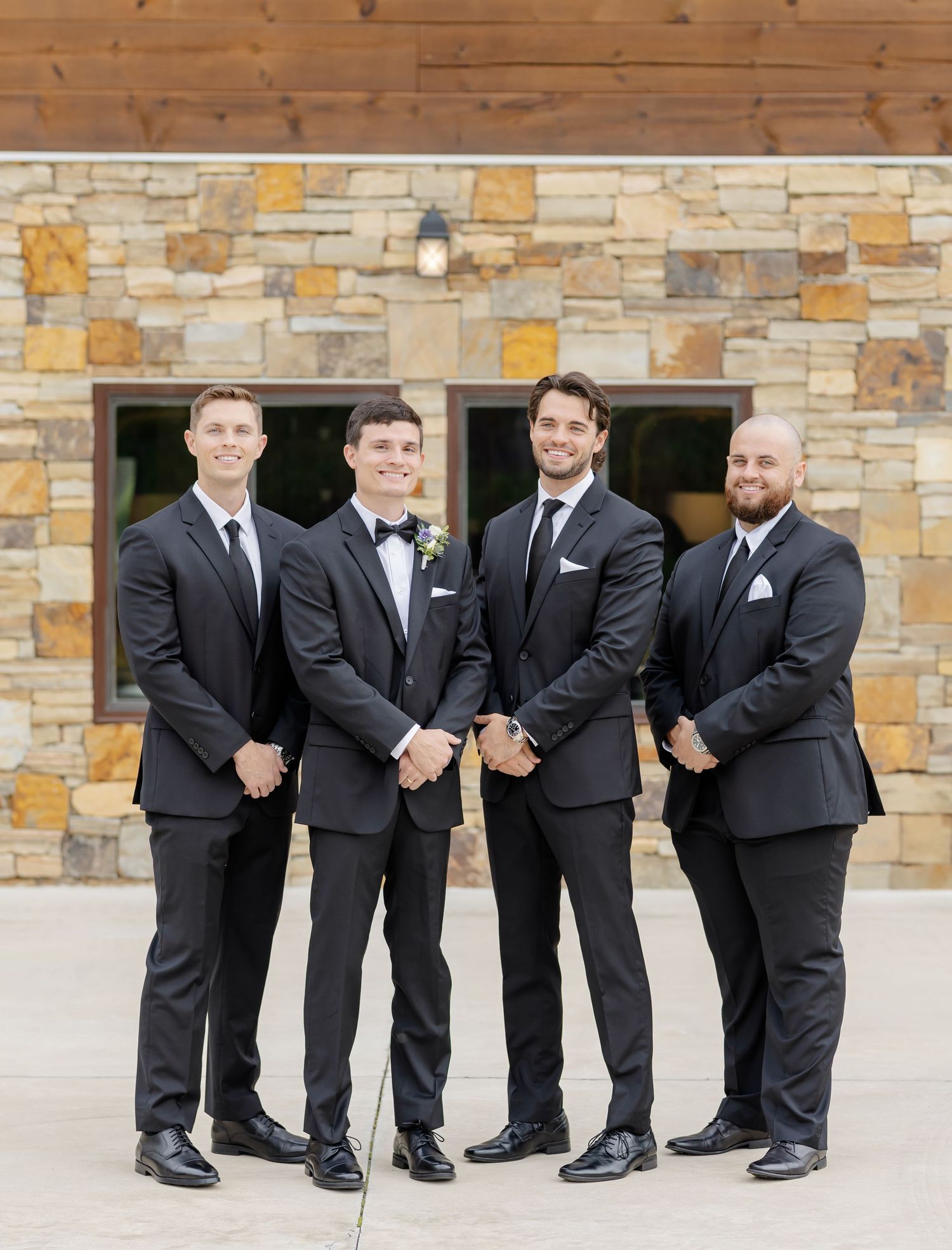 Groomsmen in black tuxedos and bow ties pose playfully in a line against a stone wall exterior.