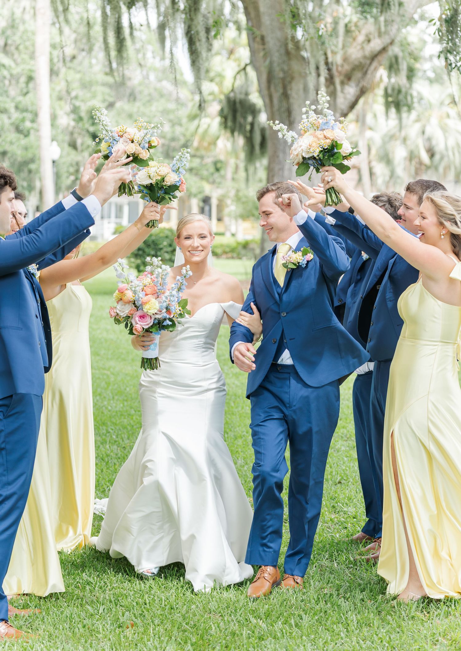 Wedding party in blue suits and yellow dresses dancing in a line under moss-draped trees in a garden setting.