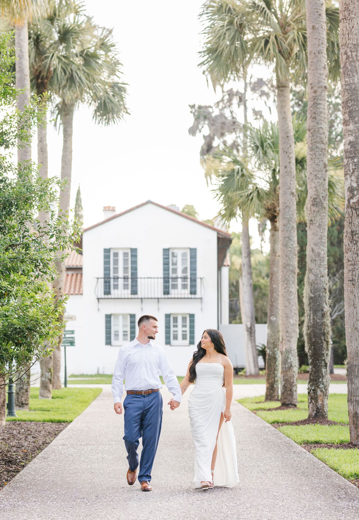 Couple walks hand-in-hand along palm-lined path toward white Southern colonial home during engagement photo session.