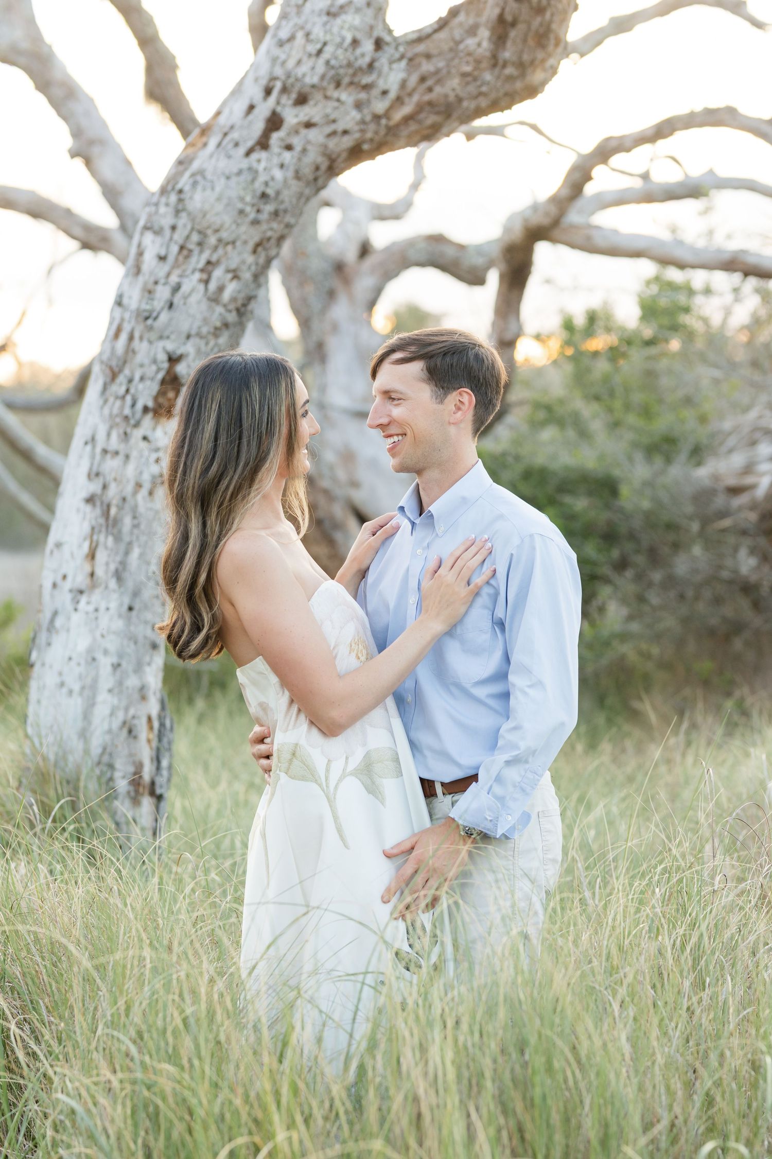 Couple sharing a romantic moment in tall grass field with a large white tree behind them.