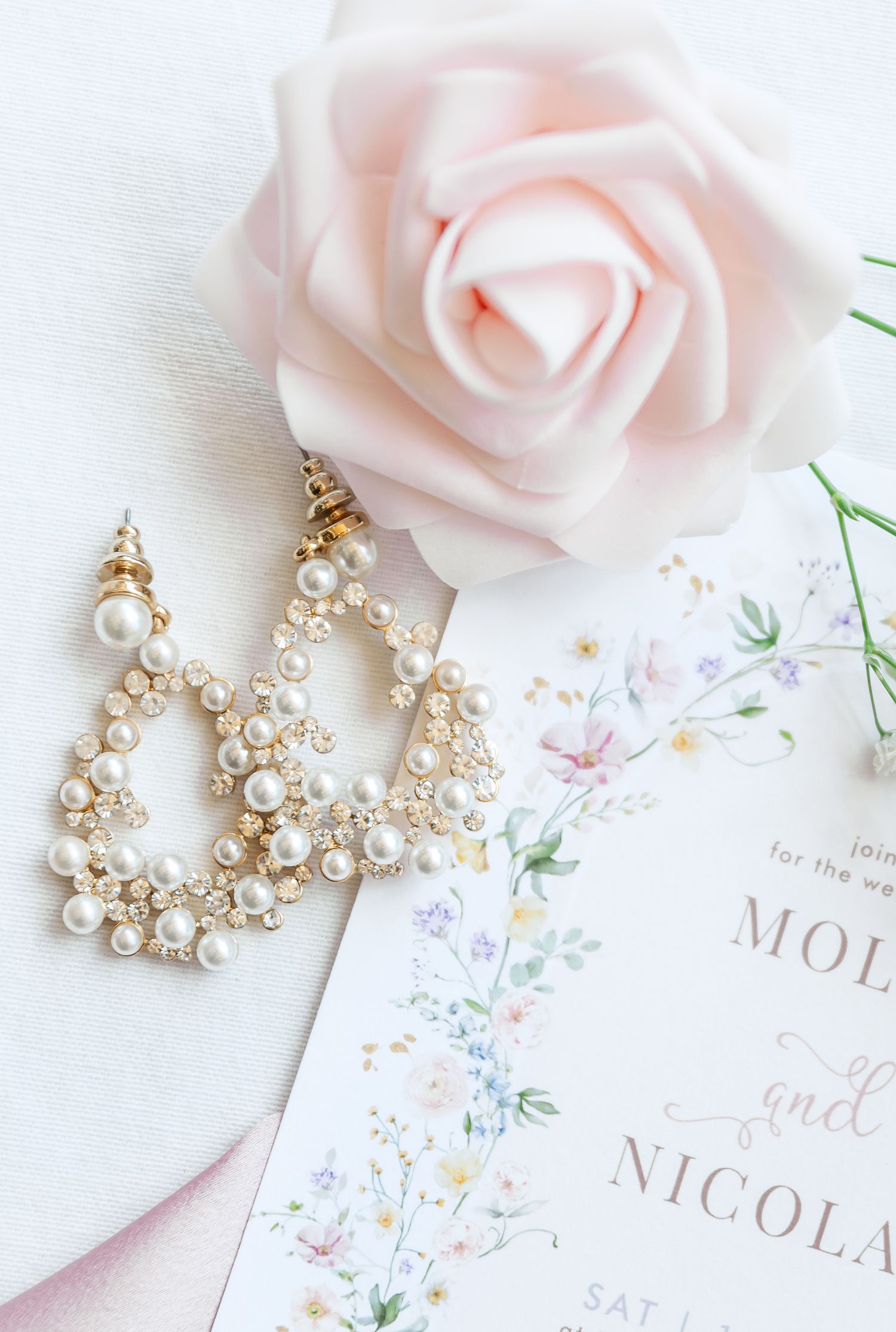 Elegant pearl and crystal wedding earrings displayed with soft pink roses and invitation cards on white background.