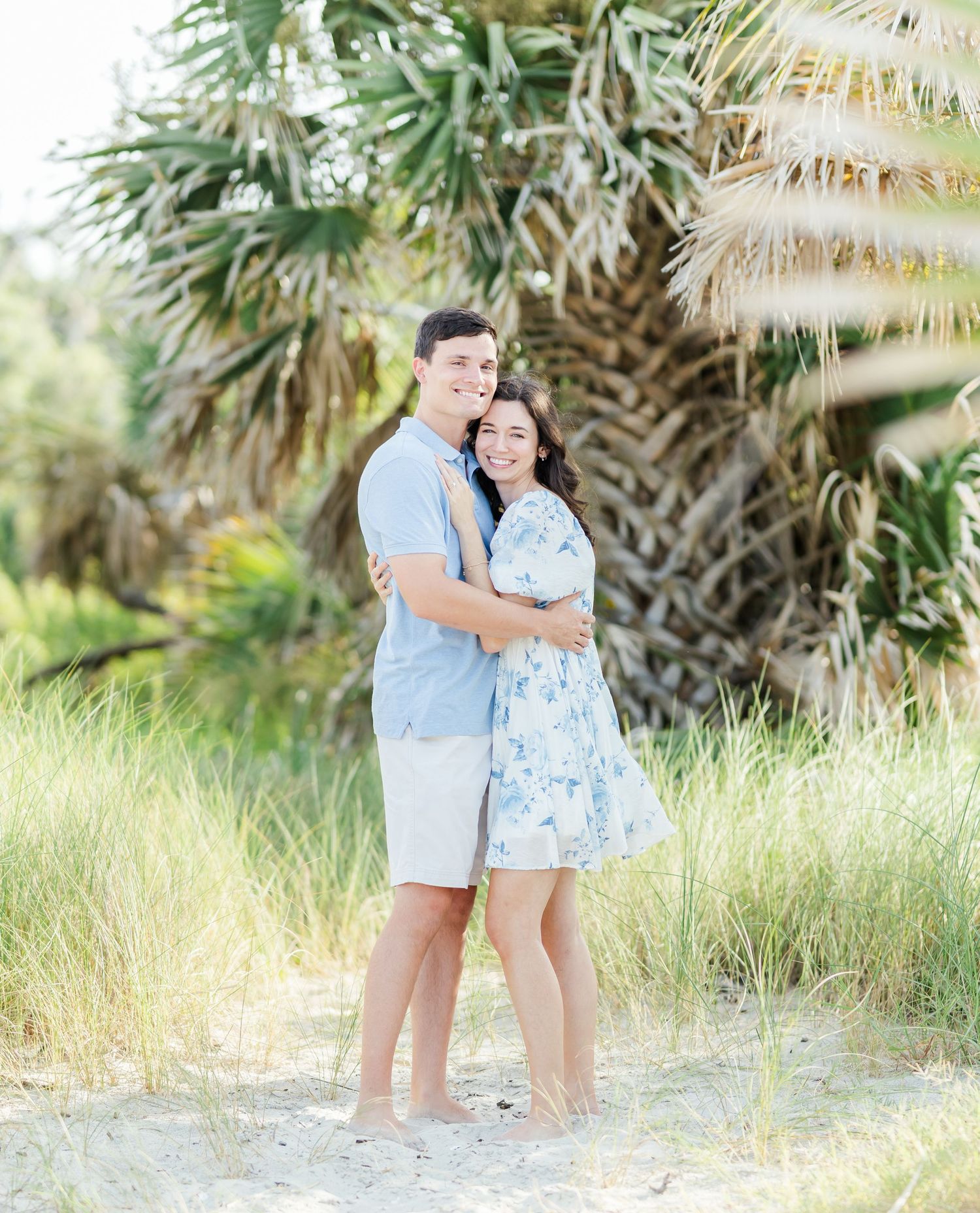 A romantic couple embraces on a beach path with tropical palm trees in the background on a sunny day.