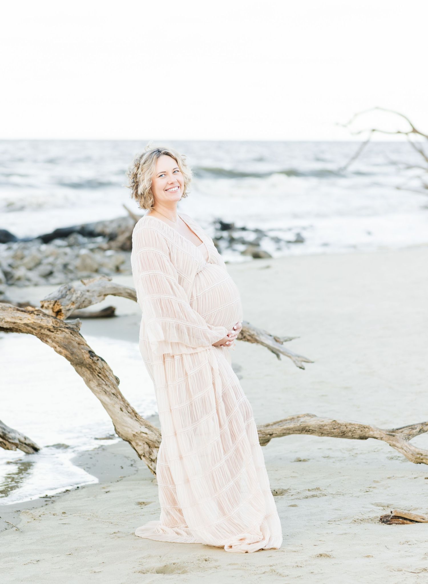 An expectant mother in a flowing blush dress poses on a driftwood log at the beach during sunset.