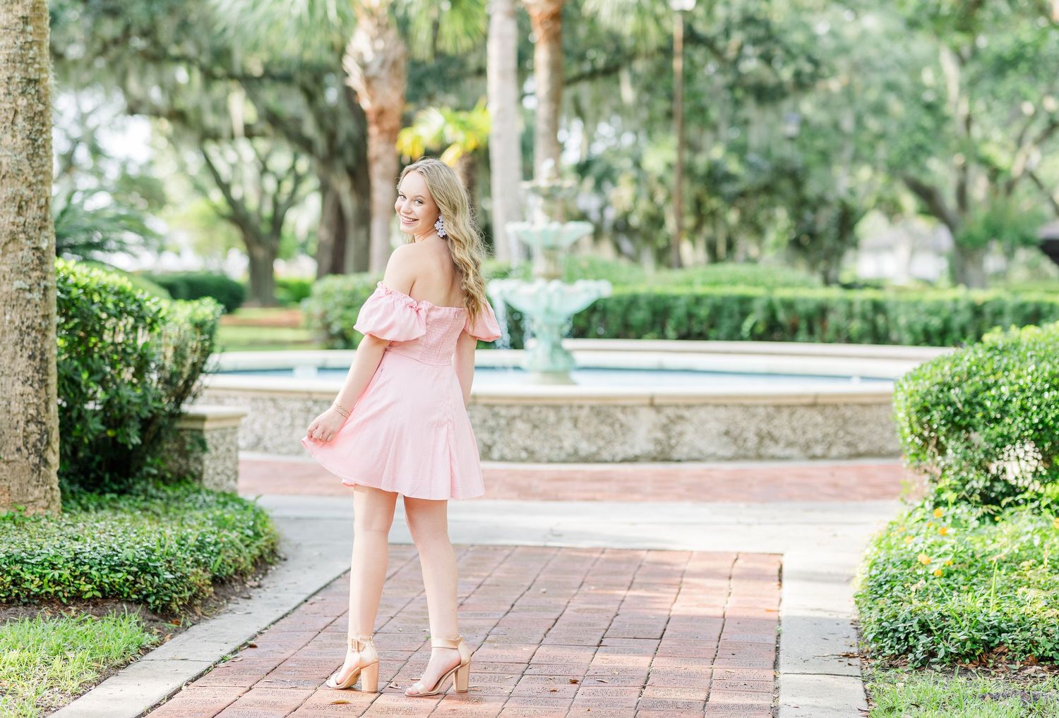 A person in a pastel pink dress glides down a brick walkway lined with lush greenery and Spanish moss.