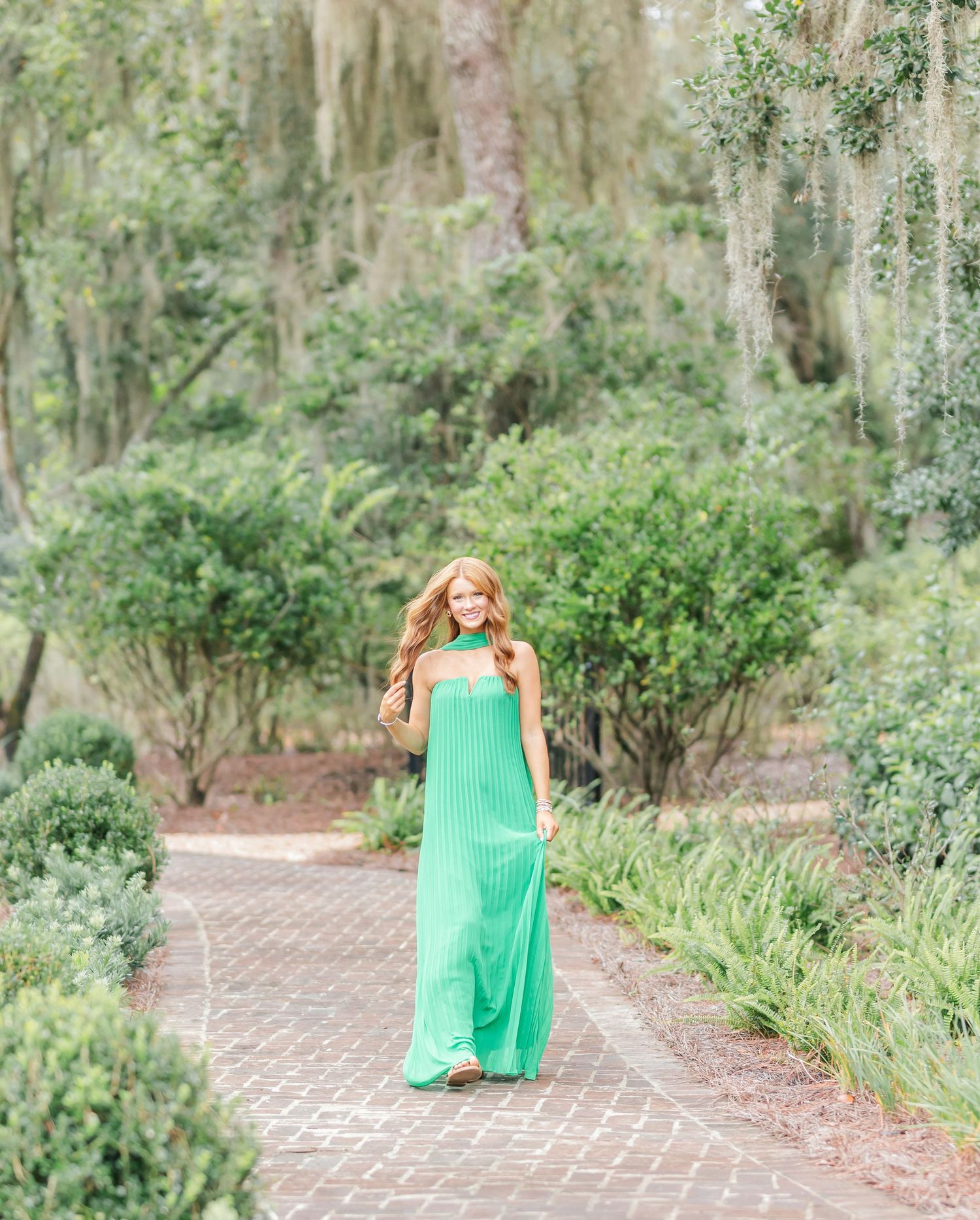 Someone in a flowing mint green maxi dress walks along a brick path surrounded by lush garden foliage and Spanish moss.