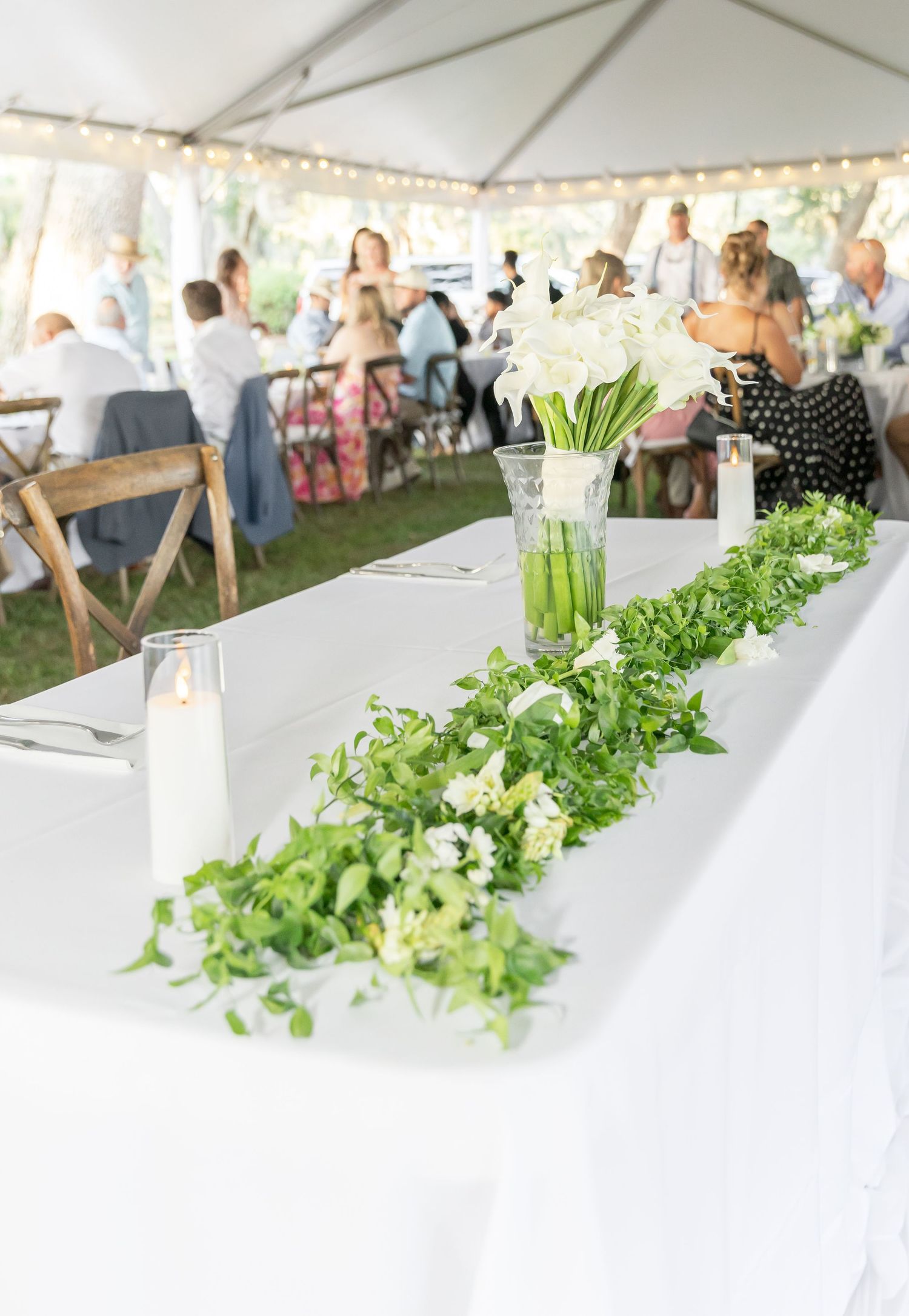White tent reception setup with elegant greenery runner and candles on dining table.