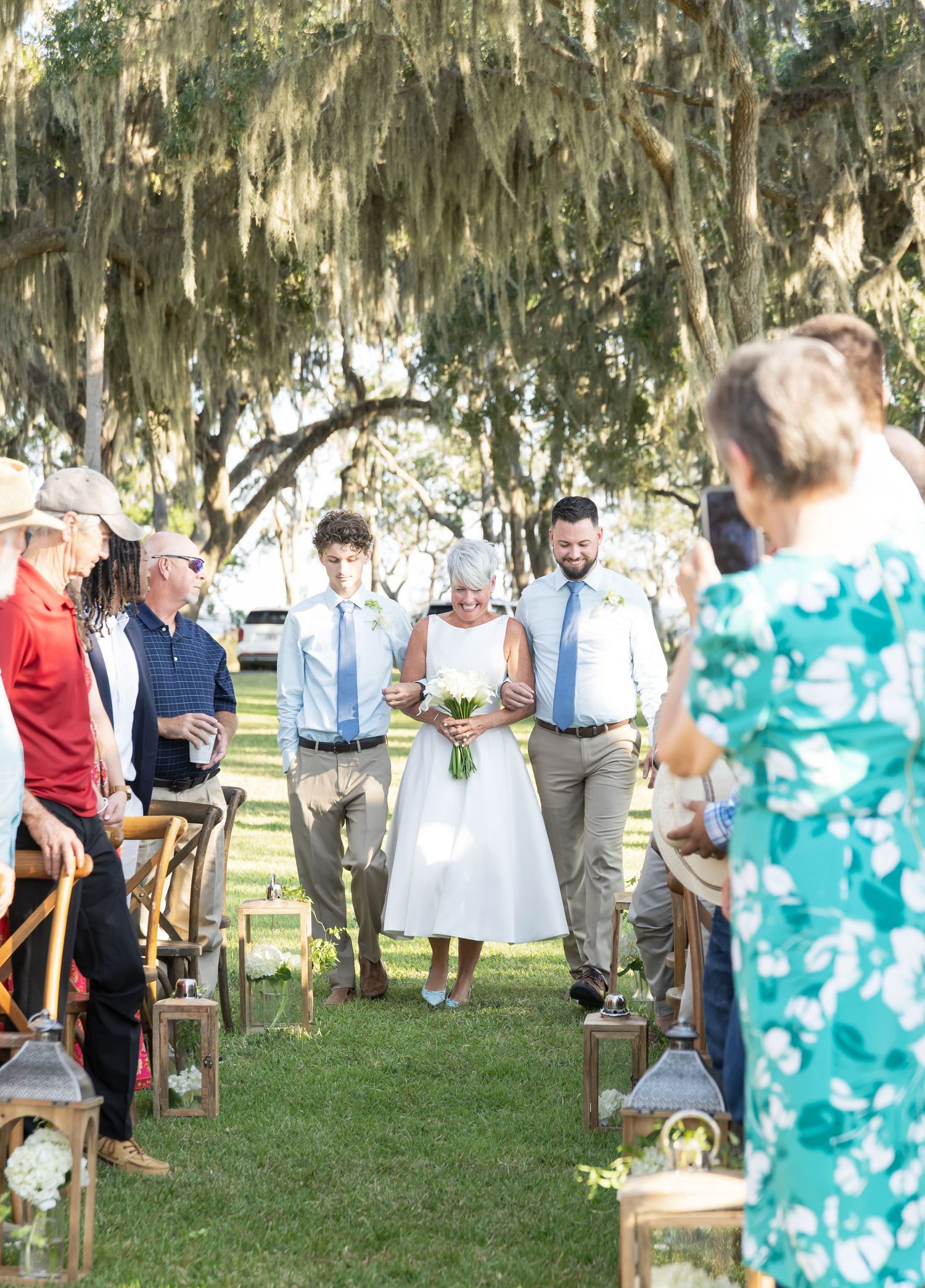 An outdoor wedding ceremony under Spanish moss-draped trees with guests seated on rustic wooden benches.