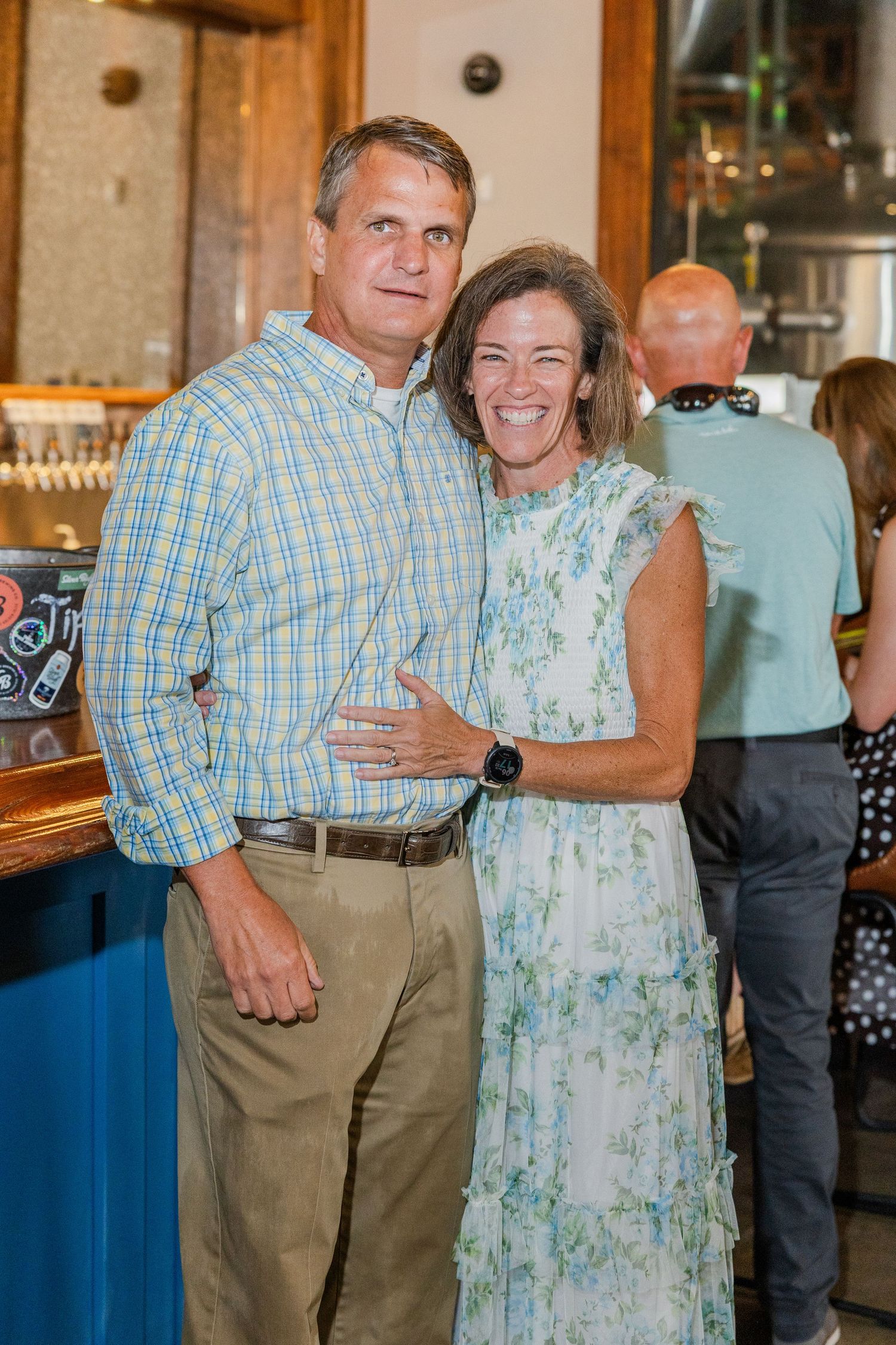 A smiling couple poses together at a casual indoor event, man in plaid shirt and woman in floral dress.
