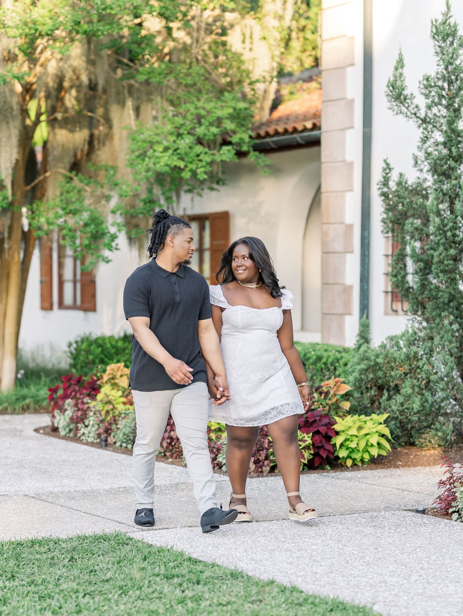 A couple holds hands and walks along a garden path outside a white building with lush greenery.