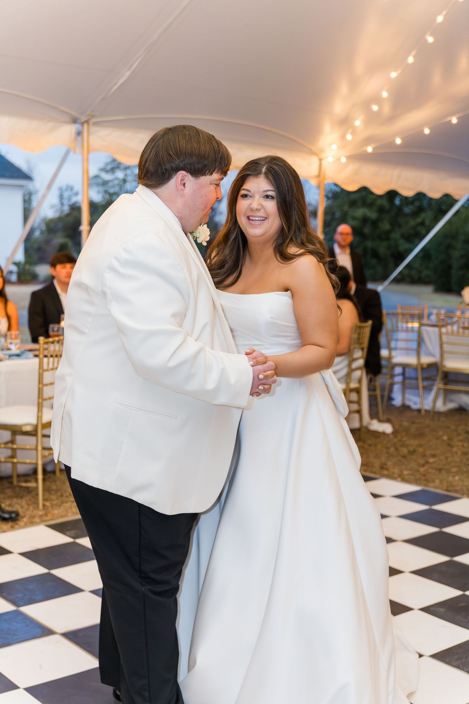 Bride and groom sharing first dance under string lights on checkered dance floor at tented reception.