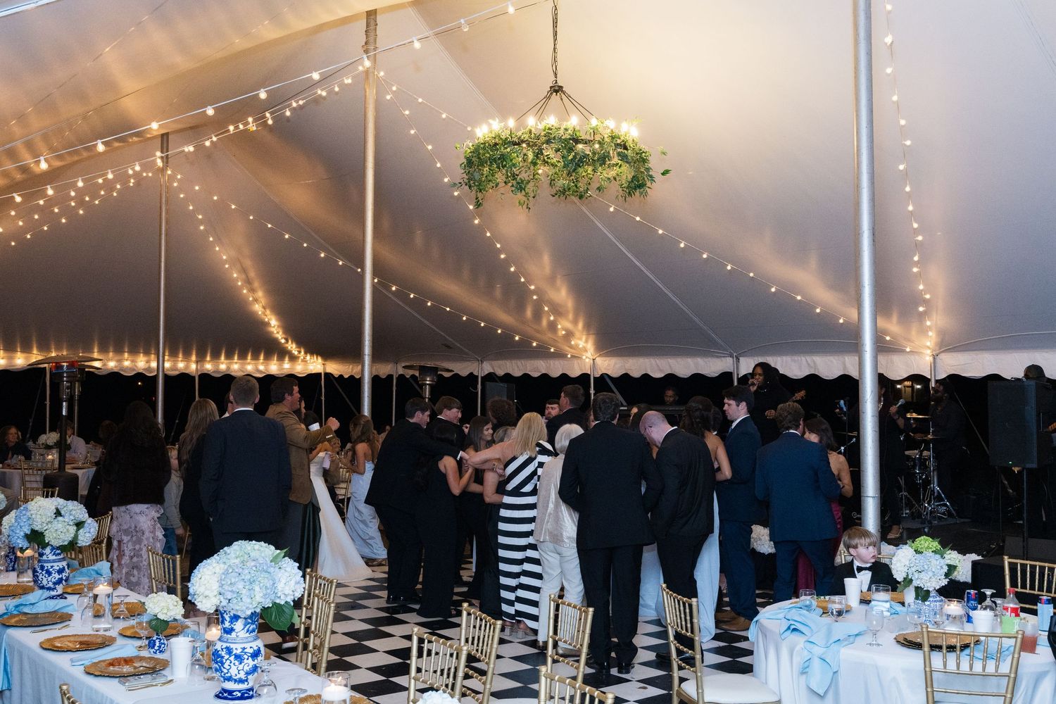 Elegant wedding reception under a white tent with string lights, black and white checkered floor, and floral chandelier.