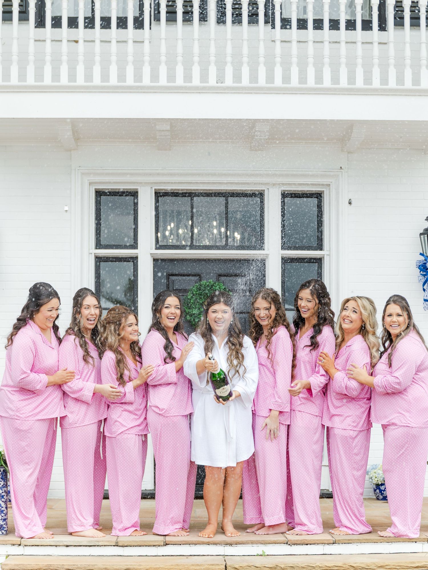 Bride in white robe pops champagne with bridesmaids in matching pink pajamas outside white venue.