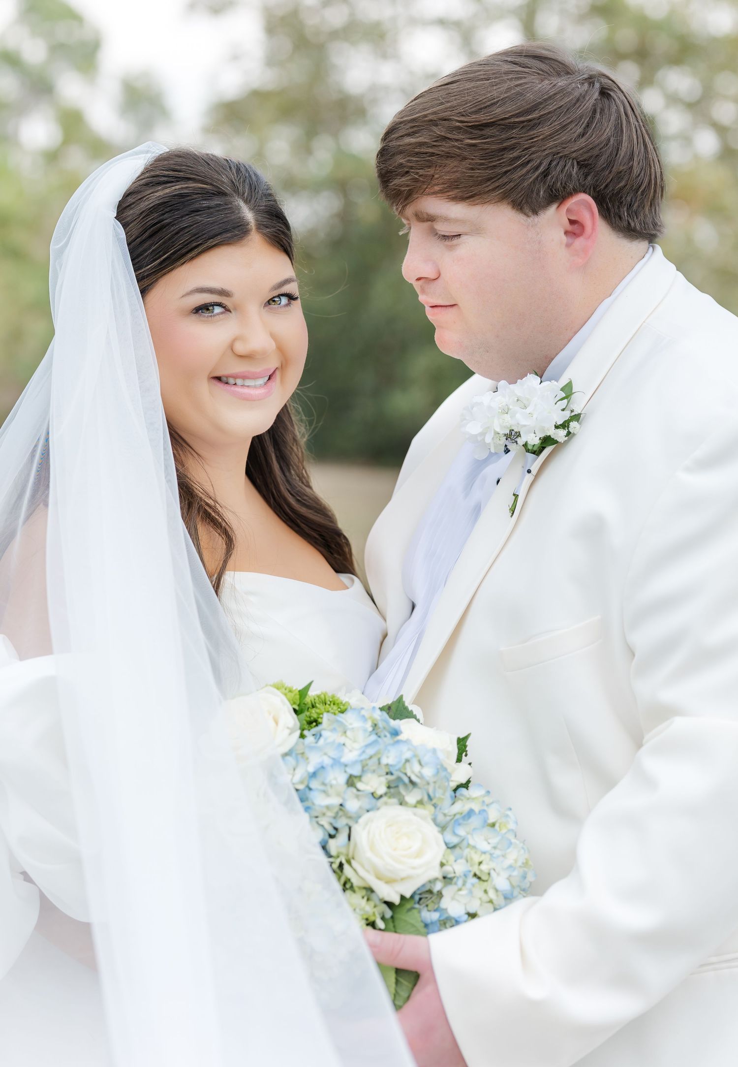 Bride in white strapless gown and veil holds blue hydrangea bouquet beside groom in white tuxedo jacket outdoors.
